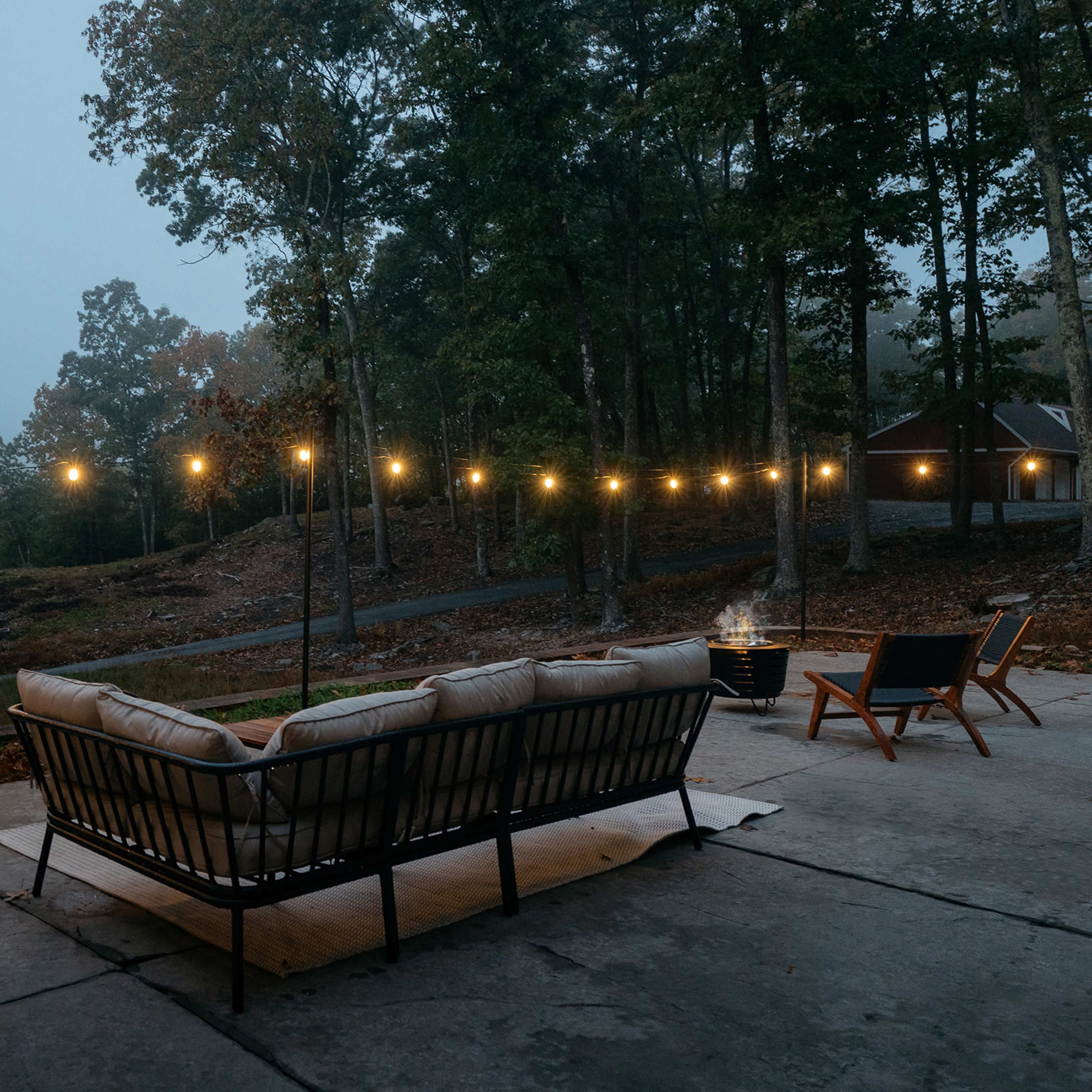 A cozy outdoor seating area with a sofa and chairs, illuminated by string lights, surrounded by trees in a misty environment.