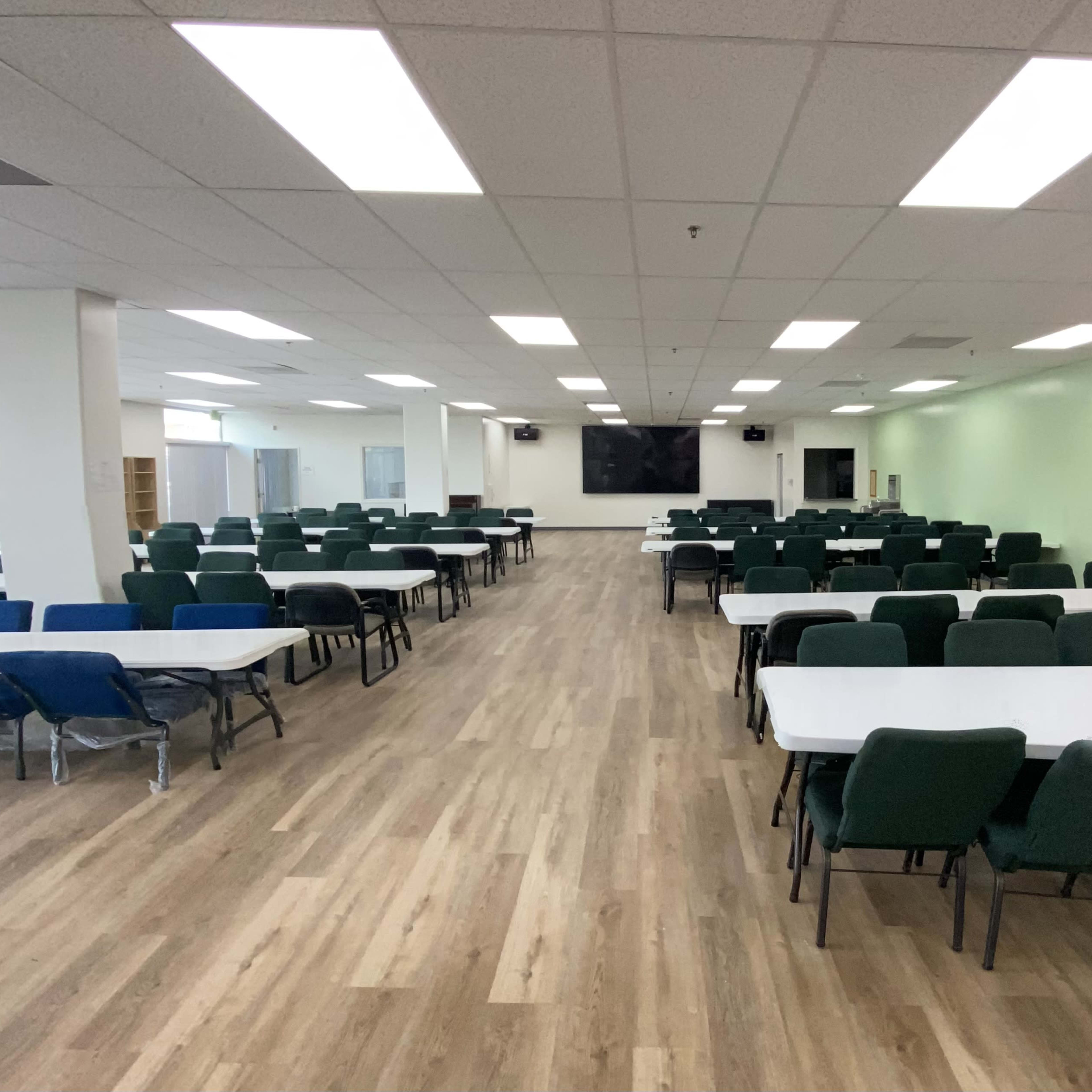 A large room with multiple rows of tables and chairs arranged for dining or meetings, featuring bright overhead lighting and wooden flooring.