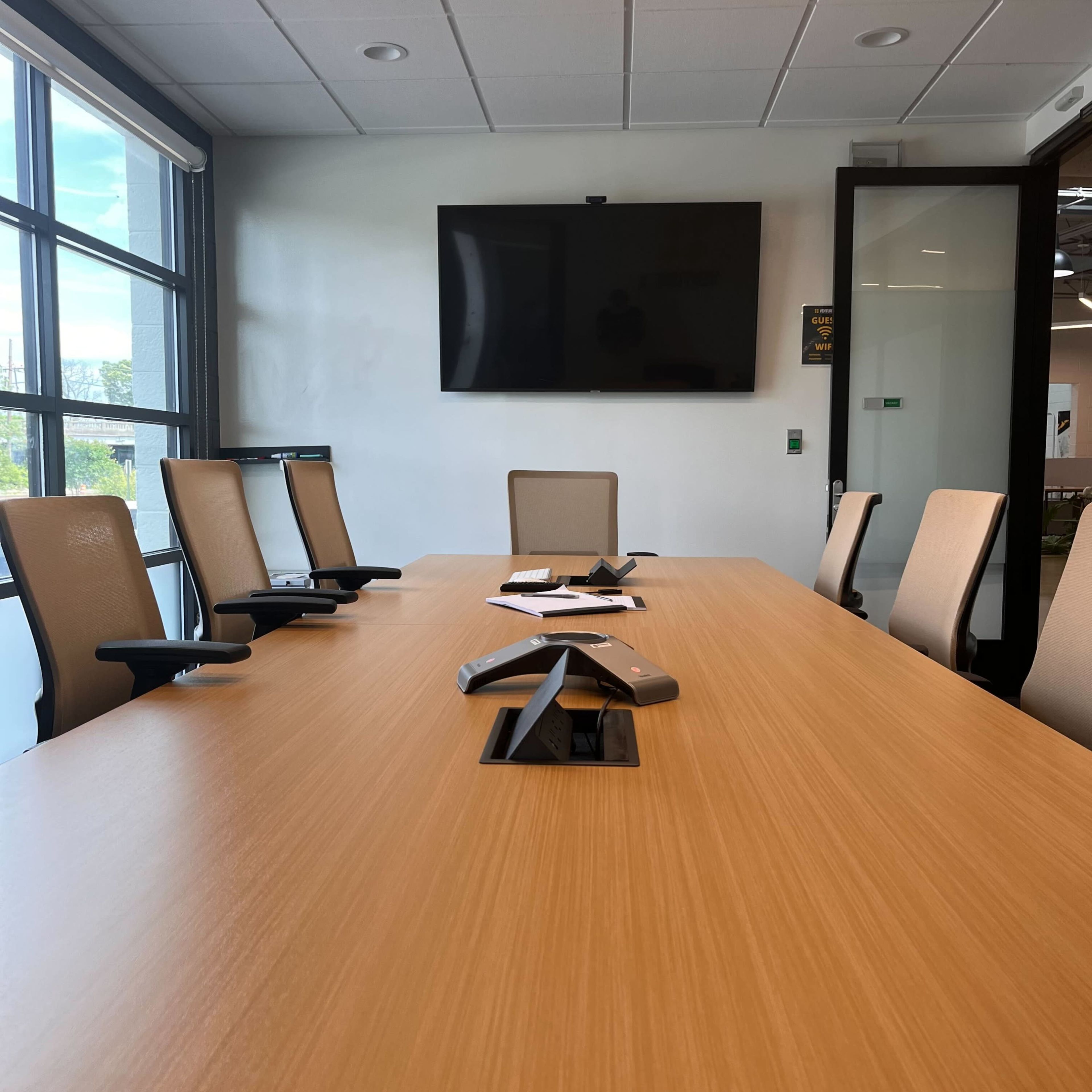 A modern conference room features a large wooden table with several chairs and a wall-mounted TV.