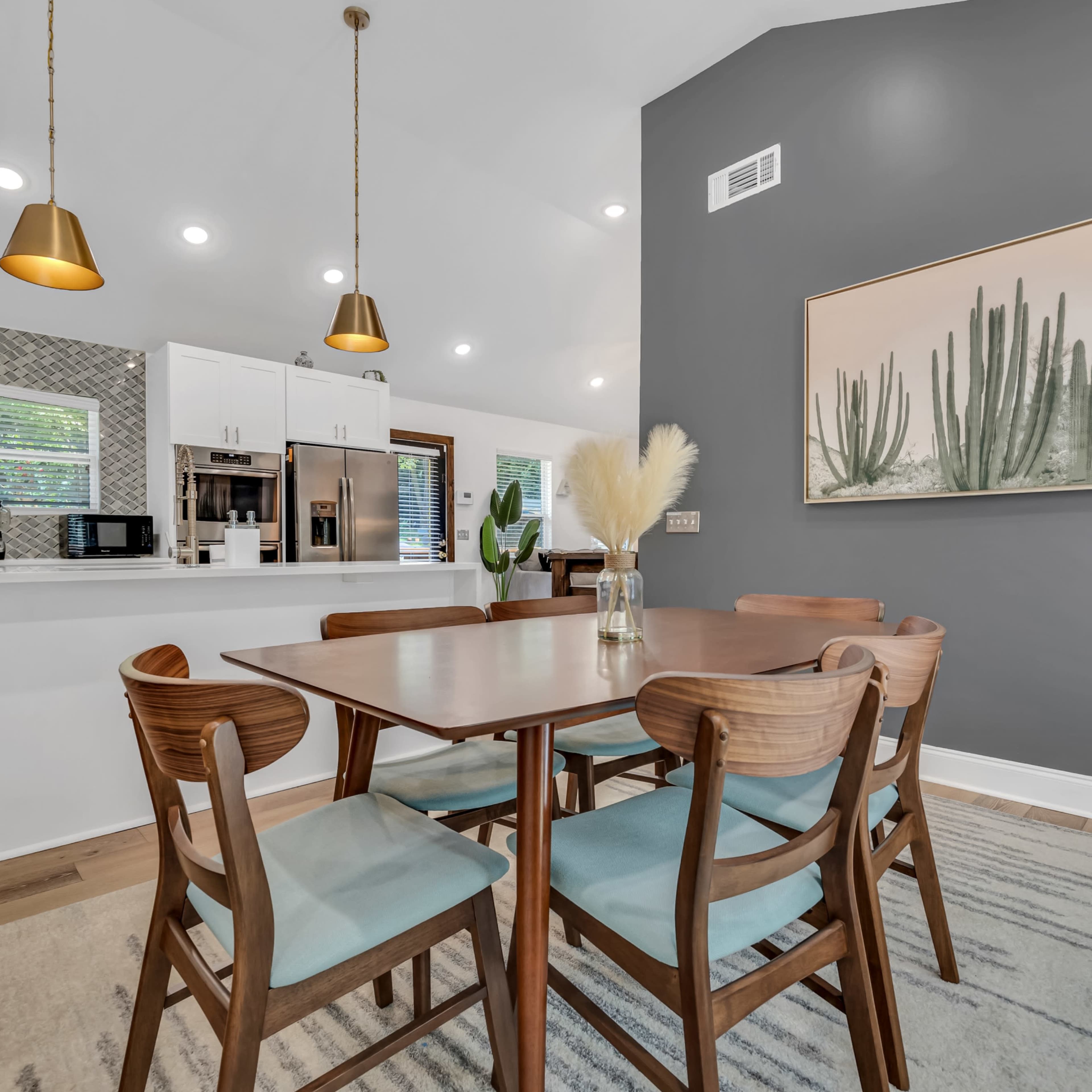 A dining area features a wooden table surrounded by six chairs, with a modern kitchen in the background and a large cactus artwork on the wall.