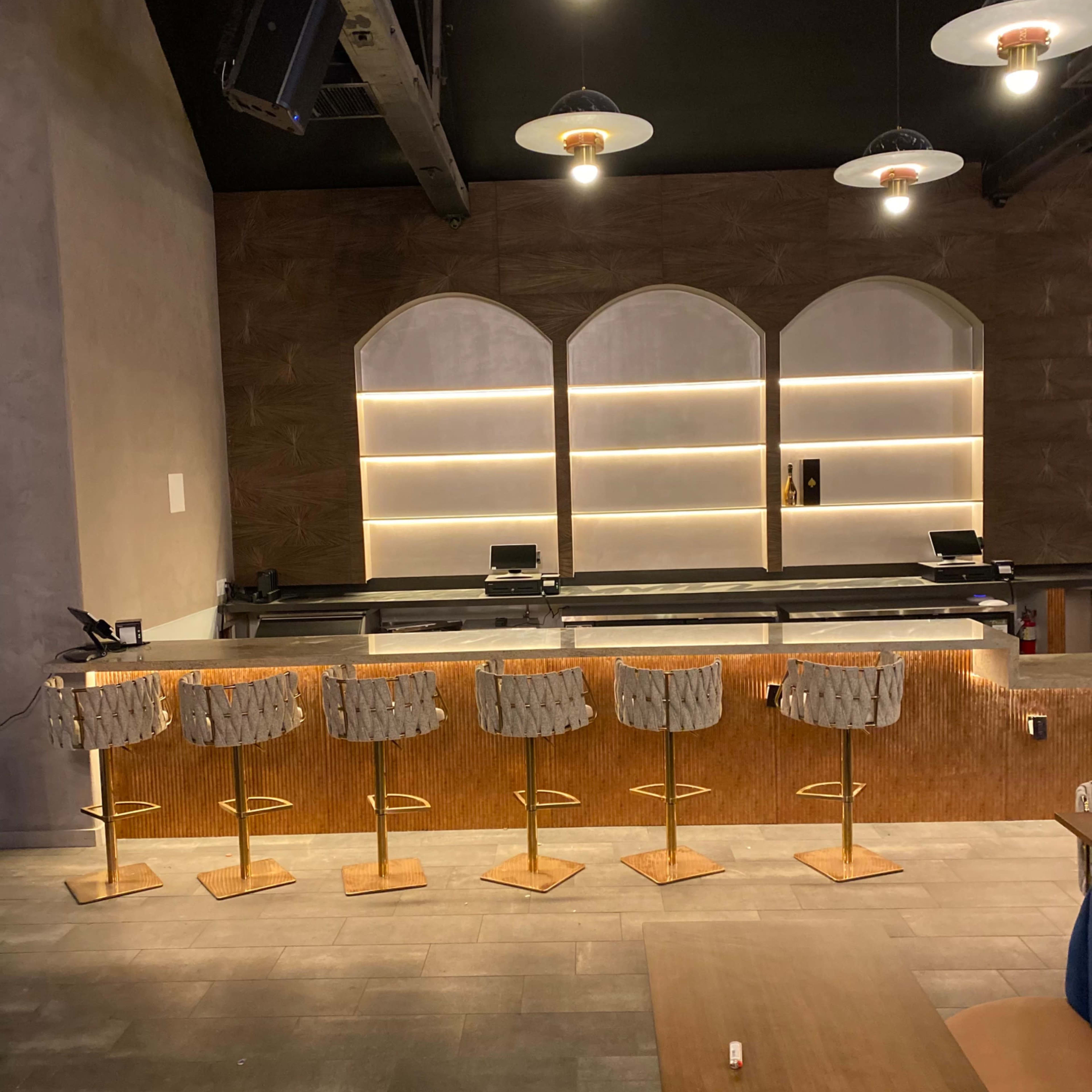 A modern bar area with a long countertop and several high stools arranged in front of it, illuminated by overhead pendant lights.