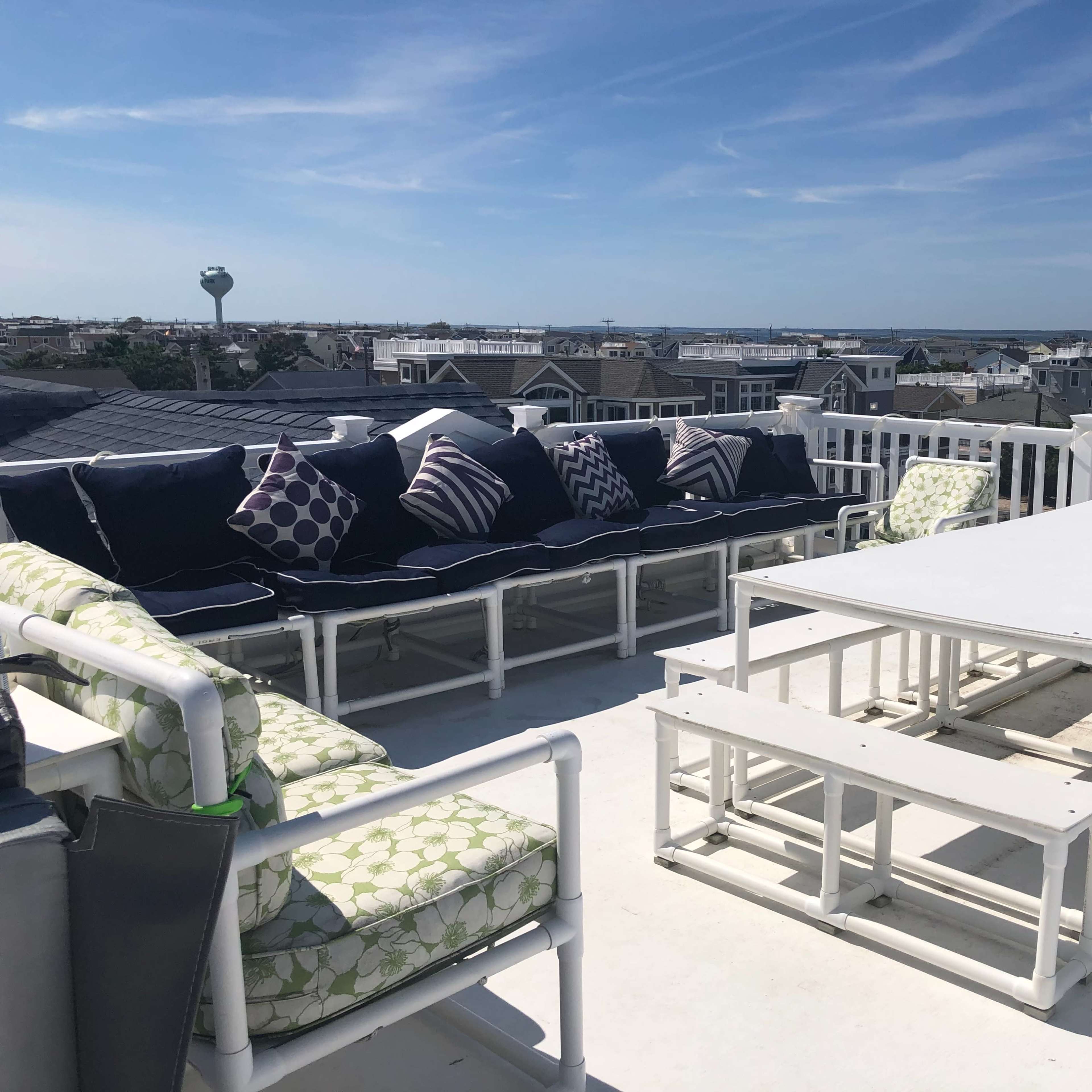 A rooftop deck with seating arrangements, including a sectional sofa and a white table, overlooking a coastal view.