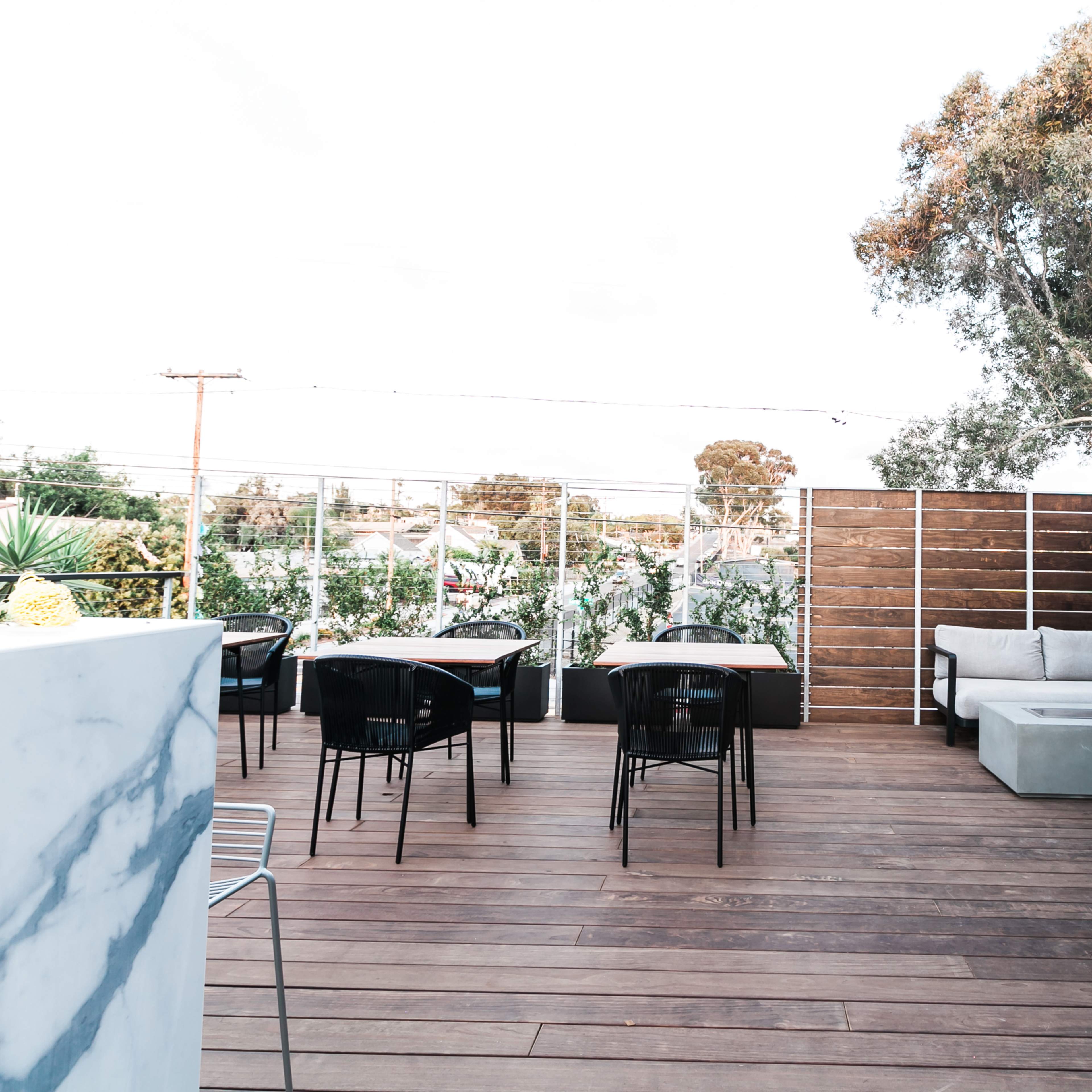 A wooden deck area furnished with tables and seating surrounded by greenery and modern fencing.