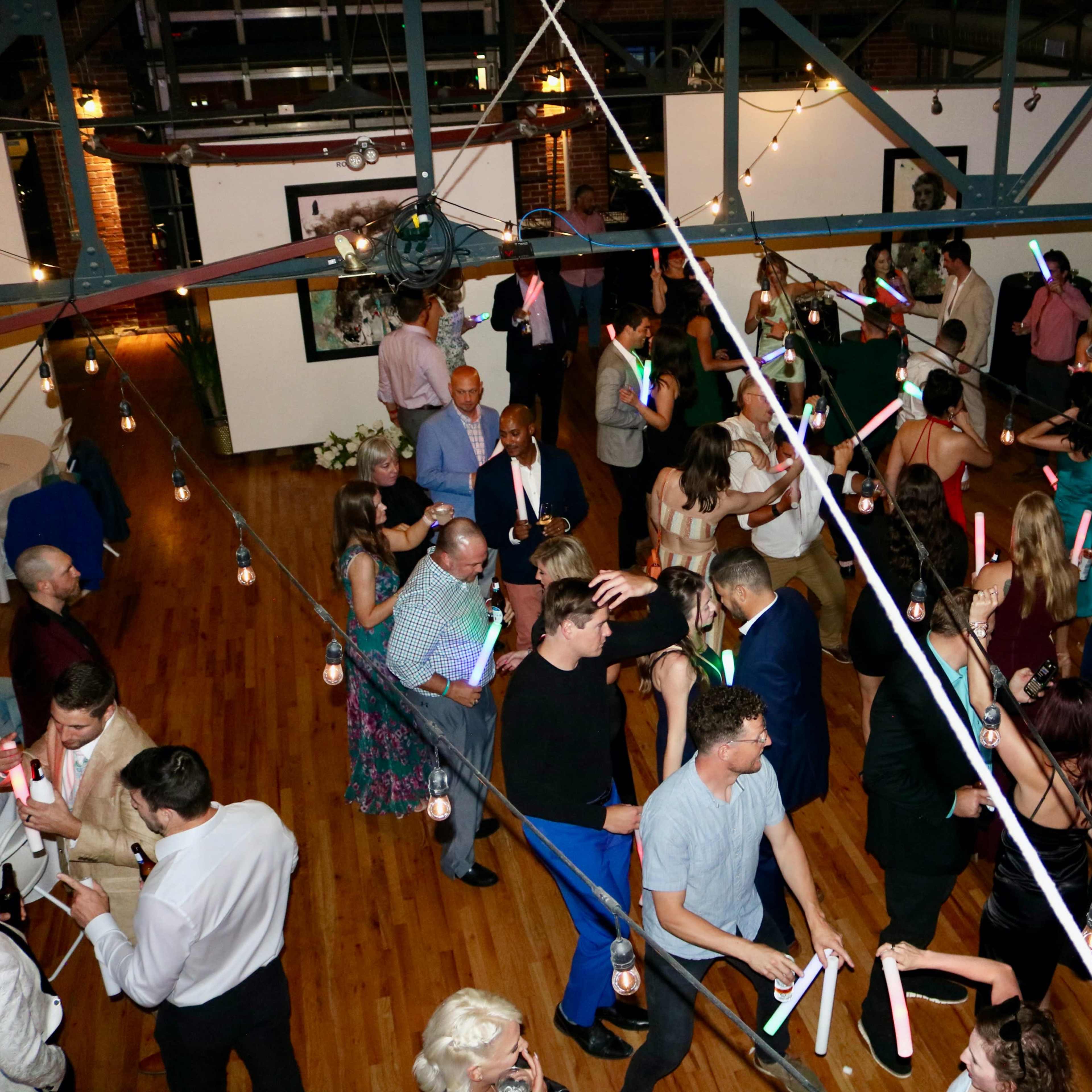 A large group of people dances with glow sticks under string lights in a spacious indoor venue.