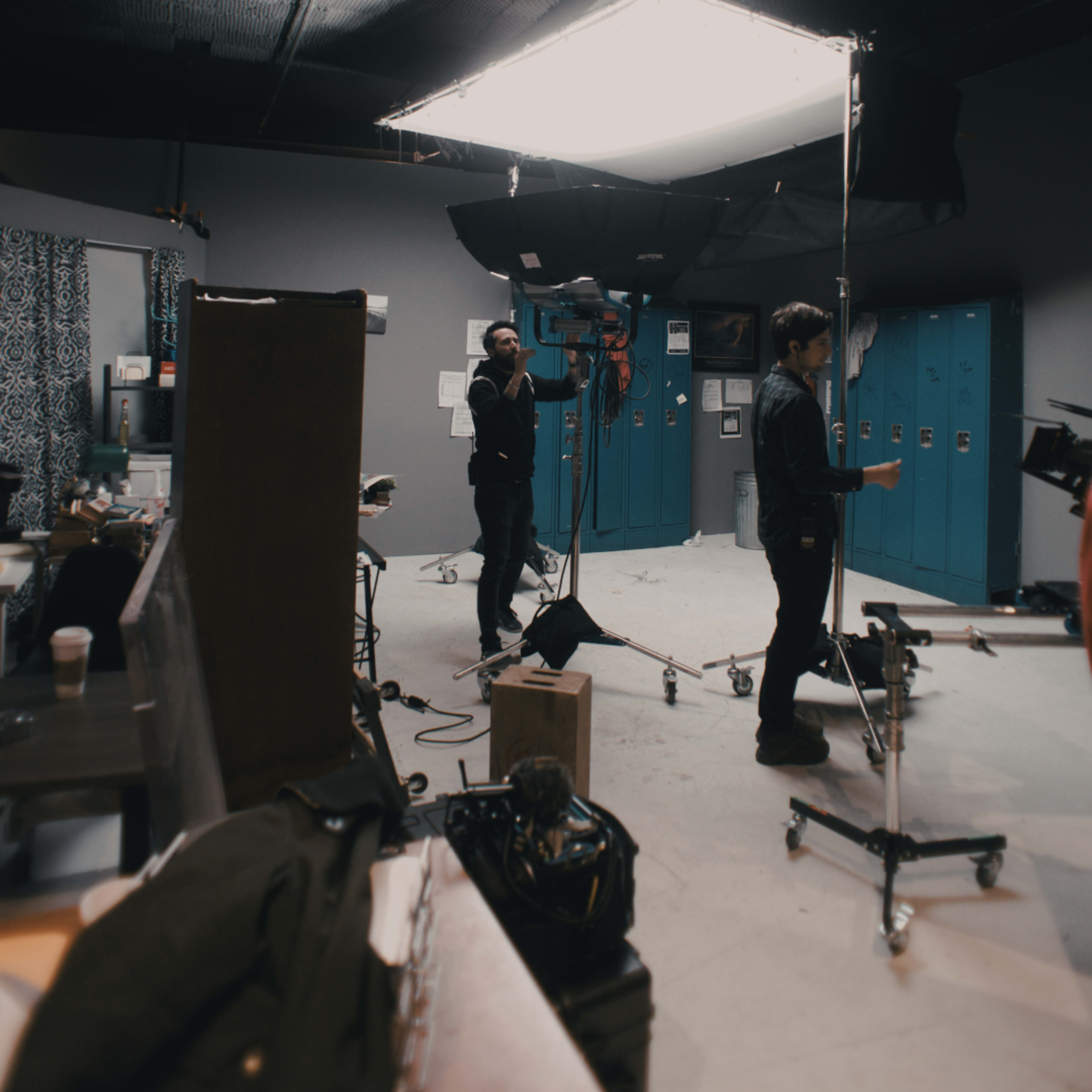 A film crew operates in a studio with blue lockers and various equipment set up for shooting.