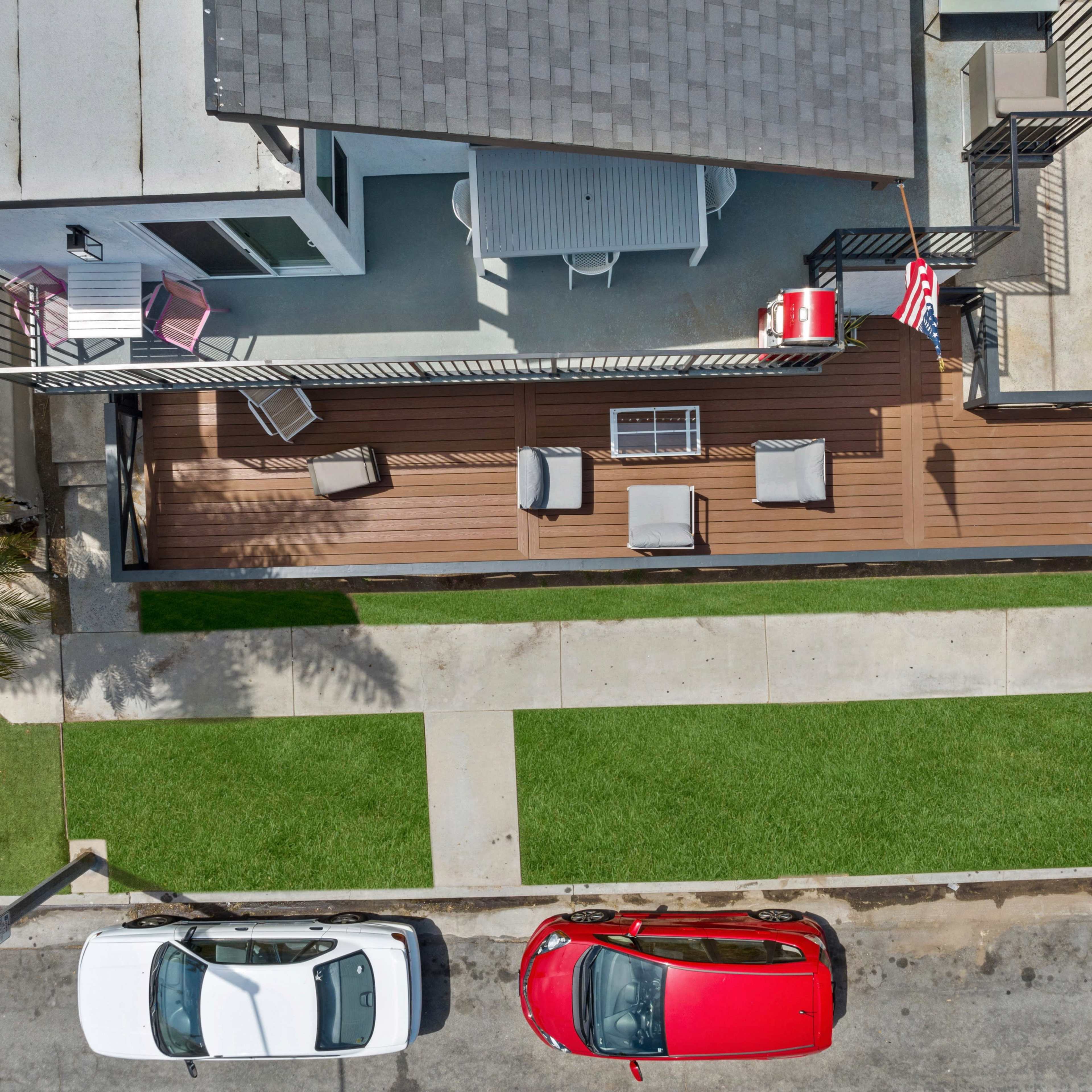 An aerial view shows a house with a wooden deck, outdoor furniture, and a small American flag, alongside a sidewalk and parked cars.