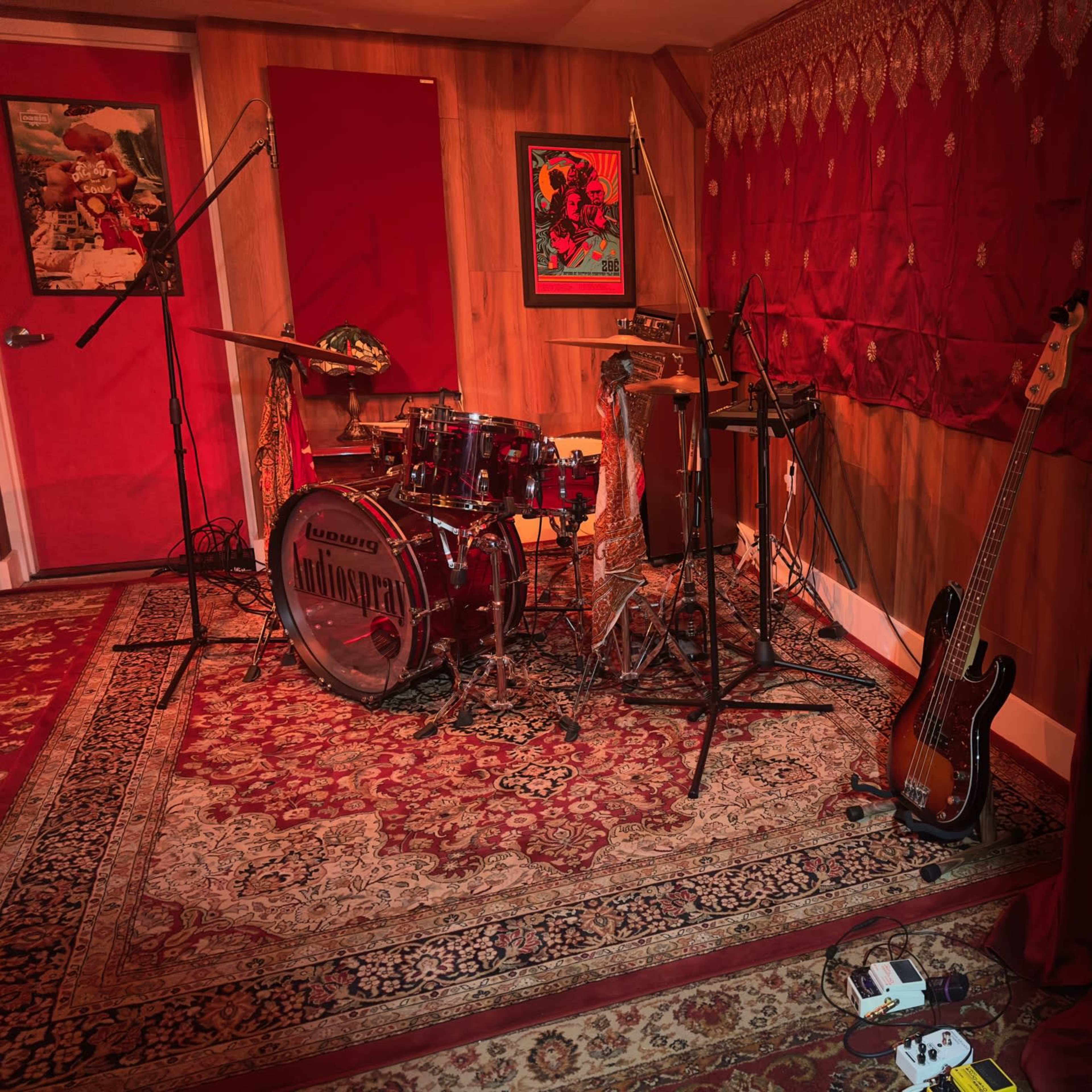 A music recording studio with a drum set, bass guitar, and various microphones arranged on red-draped walls.