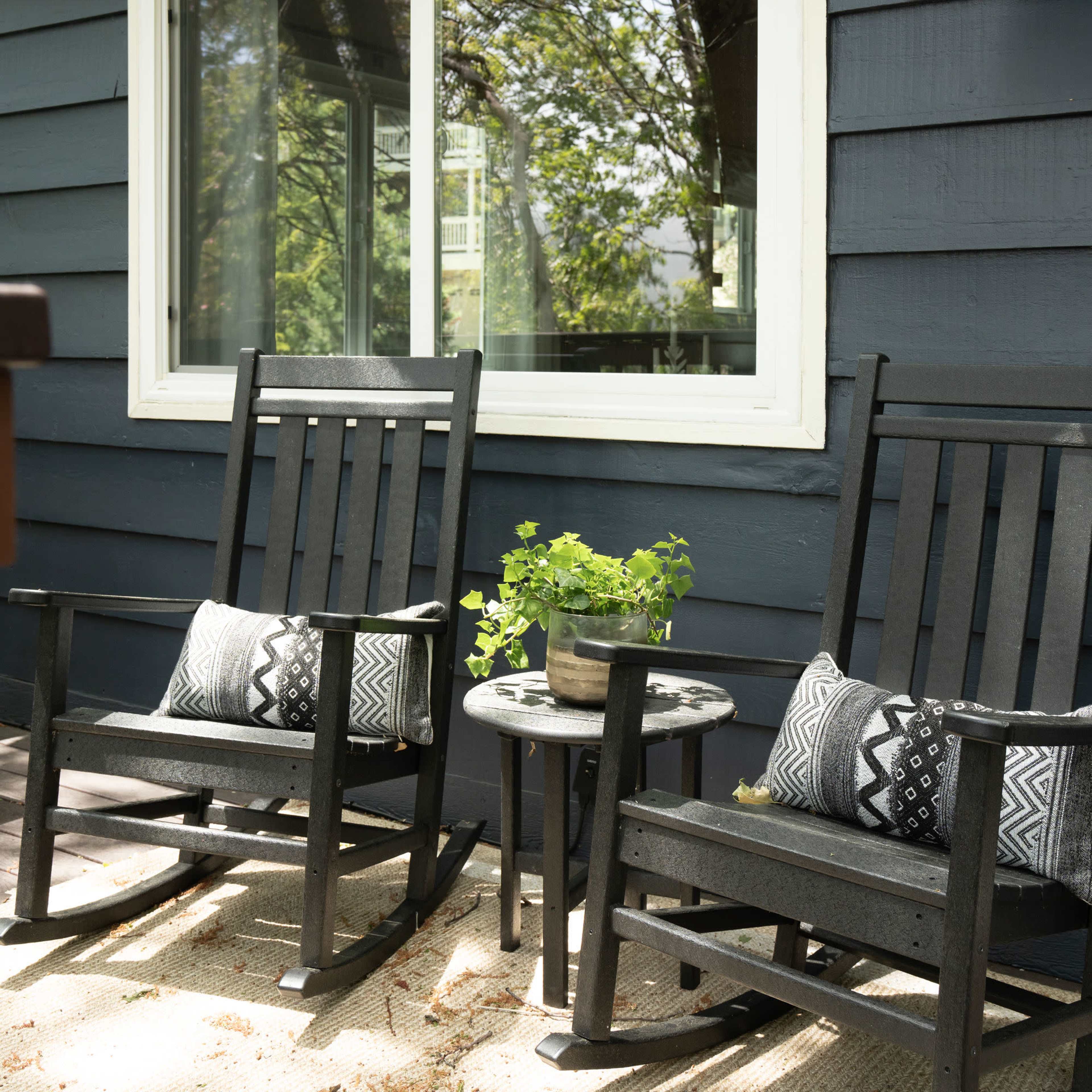 Two black rocking chairs beside a small table with a potted plant, situated in front of a window on a blue wooden house.