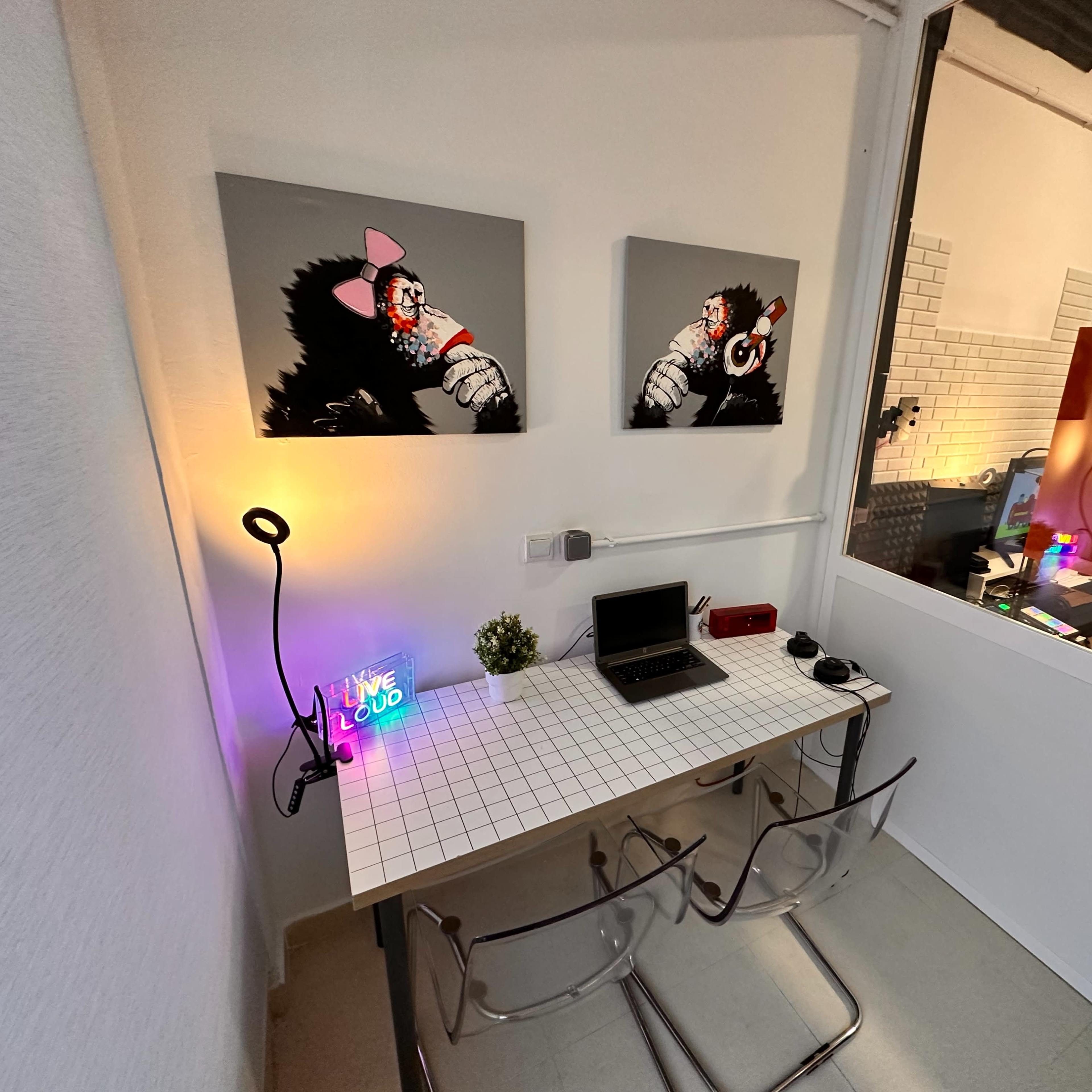 A small office space features a table with a laptop, vibrant artwork on the walls, and decorative lighting.
