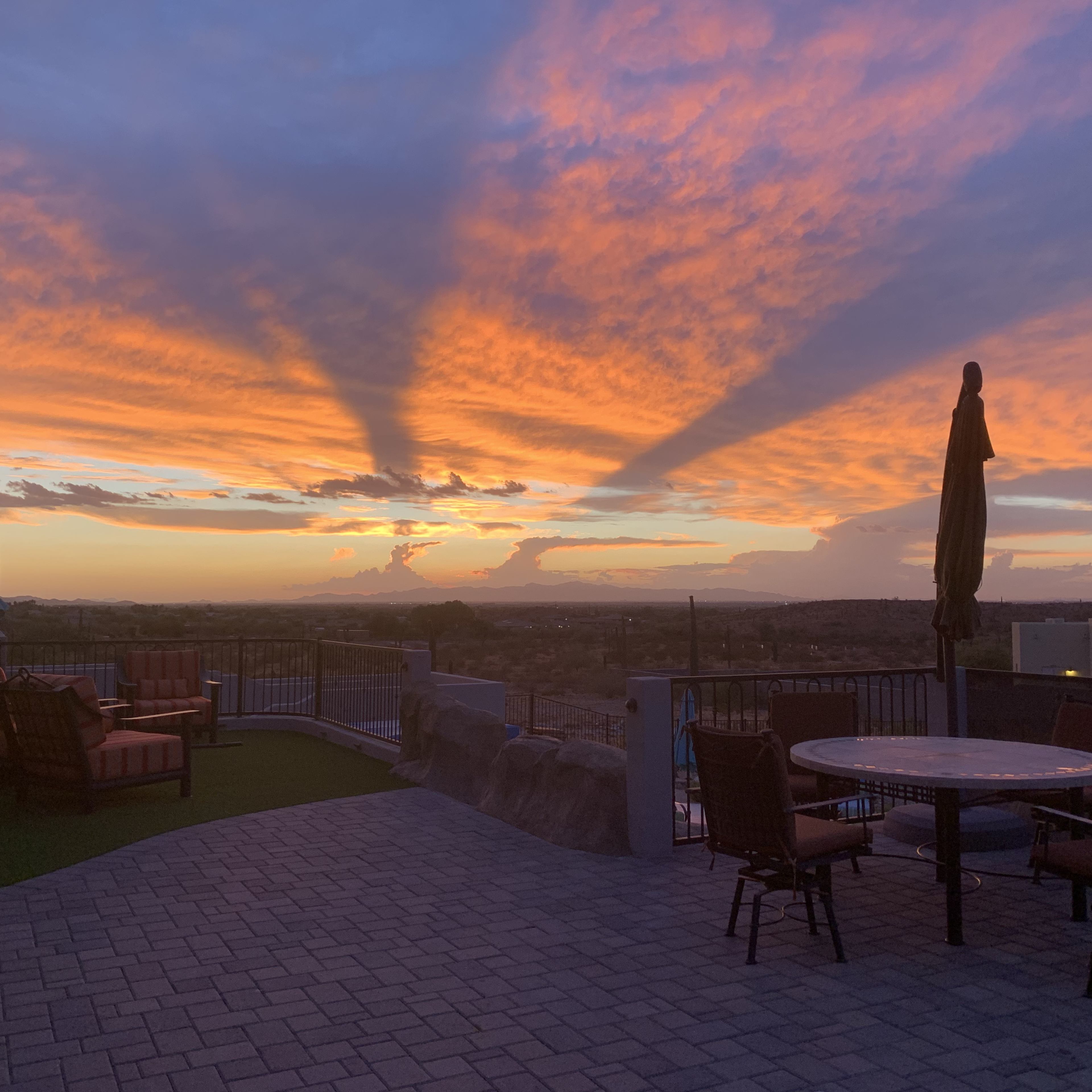 The scene shows a patio with furniture overlooking a vibrant sunset with long shadows stretching across the sky.