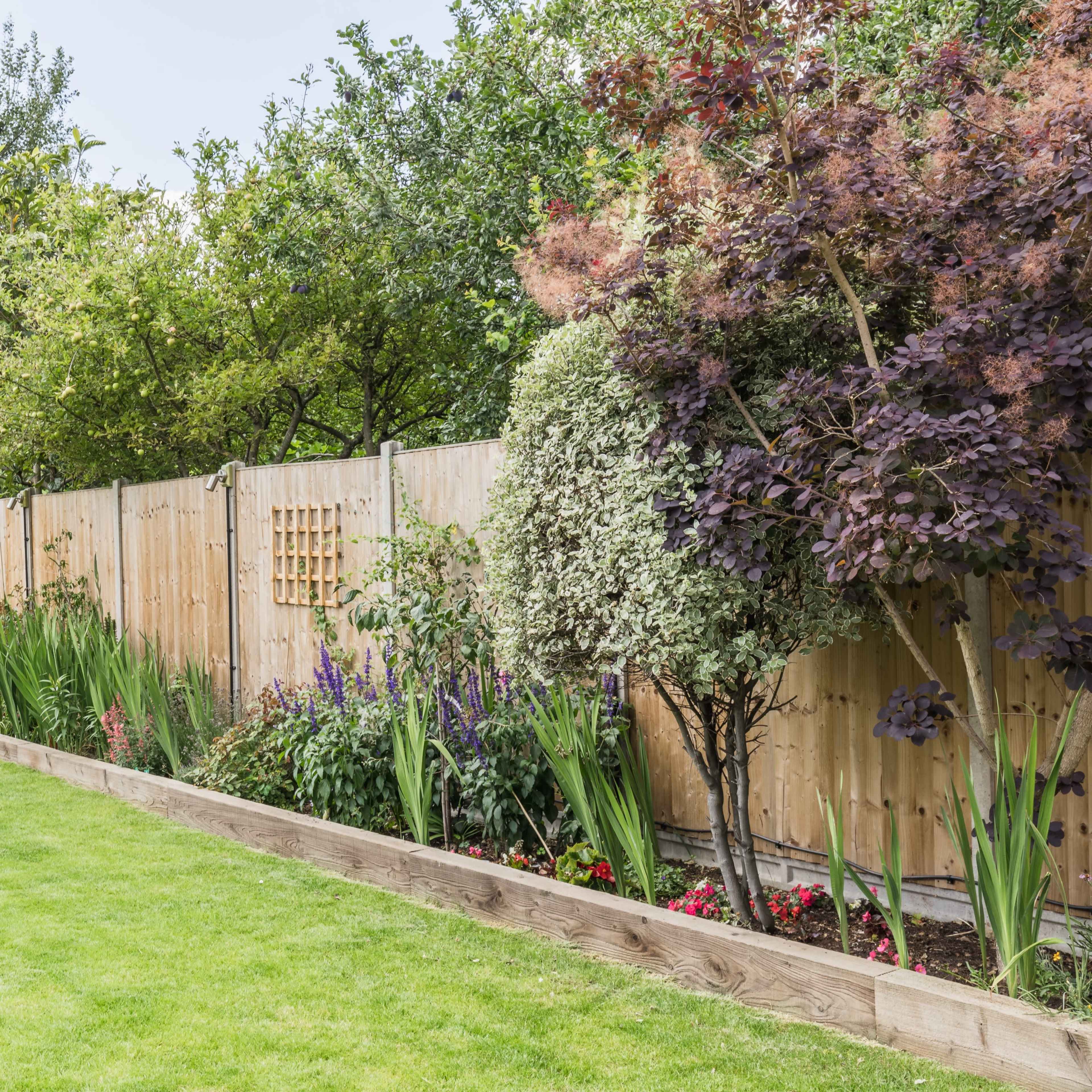 A well-kept garden features neatly arranged flower beds along a wooden fence, with two white deck chairs set on the green lawn.