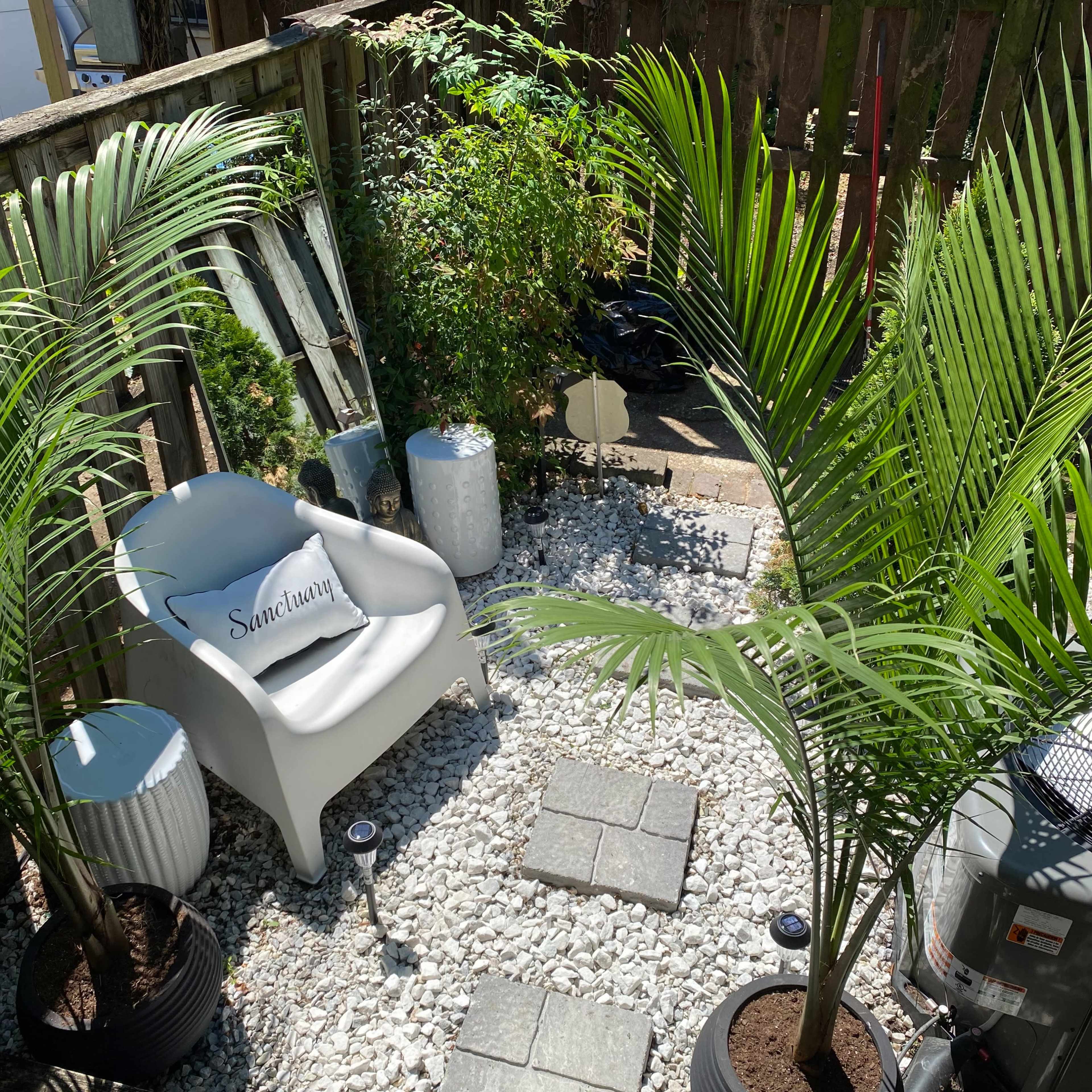 A small outdoor space with a white chair, two decorative pots, stones, and palm plants, arranged around a pathway of gray stepping stones.