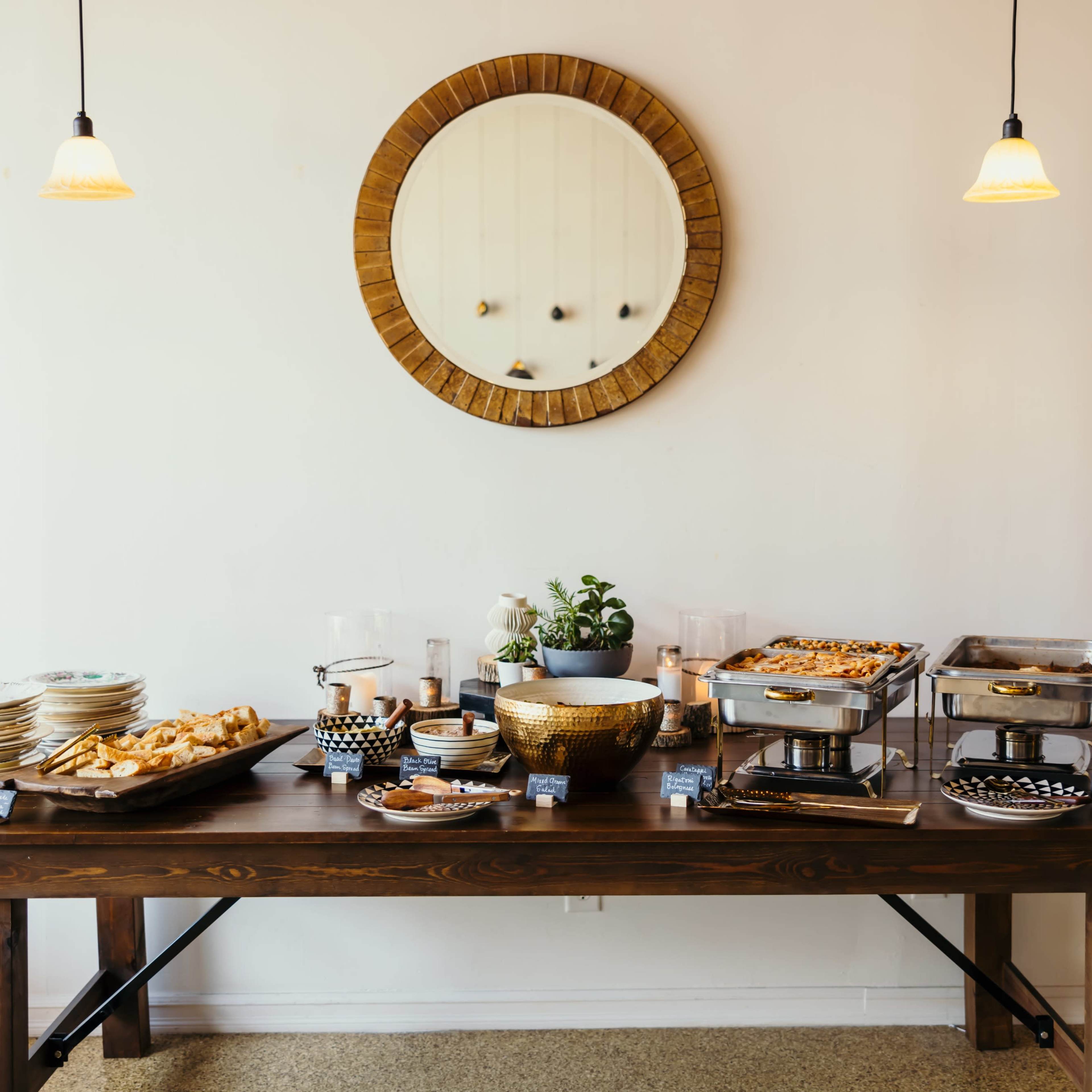 A long wooden table is set with a variety of food dishes, plates, and utensils against a plain wall featuring a round mirror and two hanging lights.