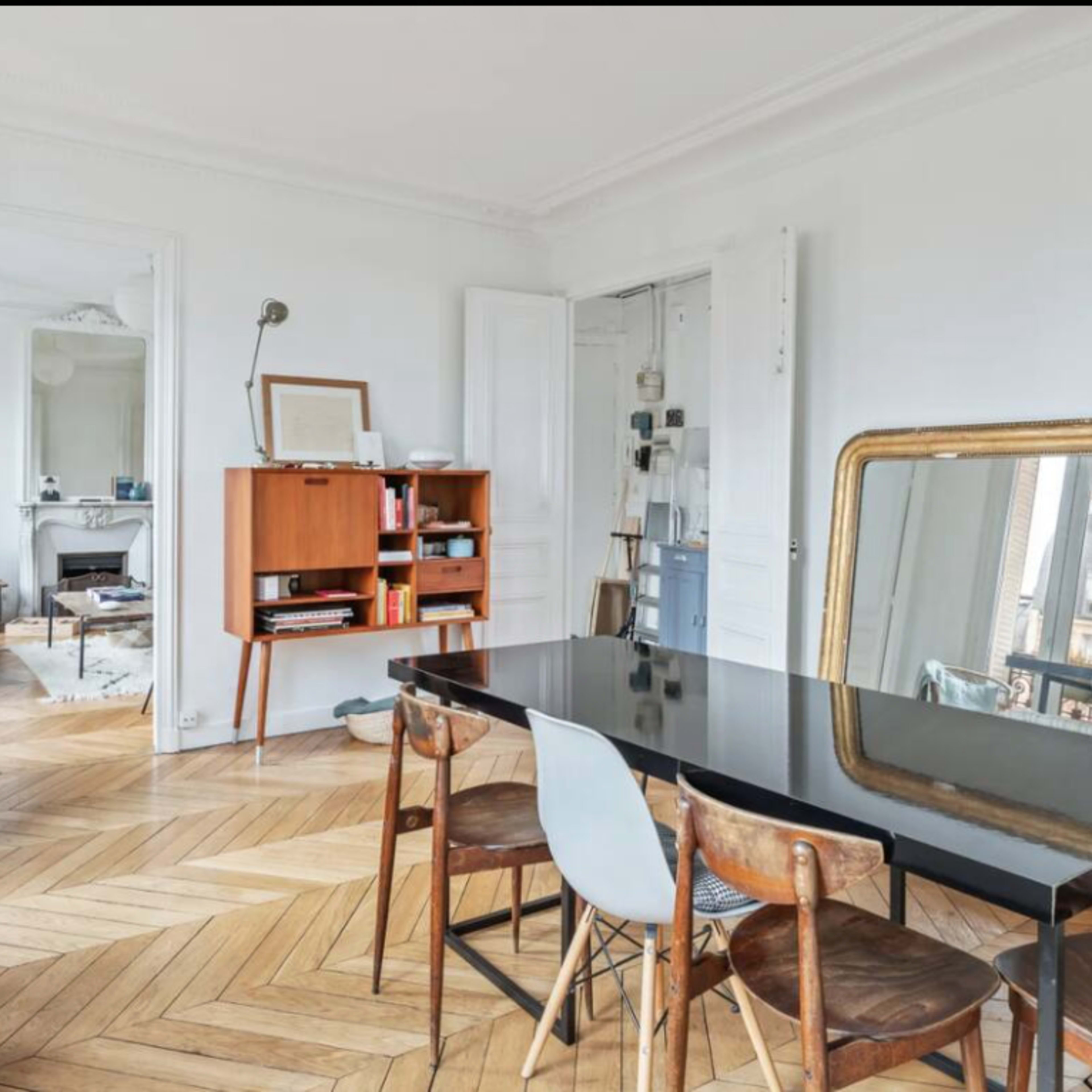 A modern living space with a black dining table surrounded by wooden chairs, a minimalist bookshelf, and large windows that provide natural light.