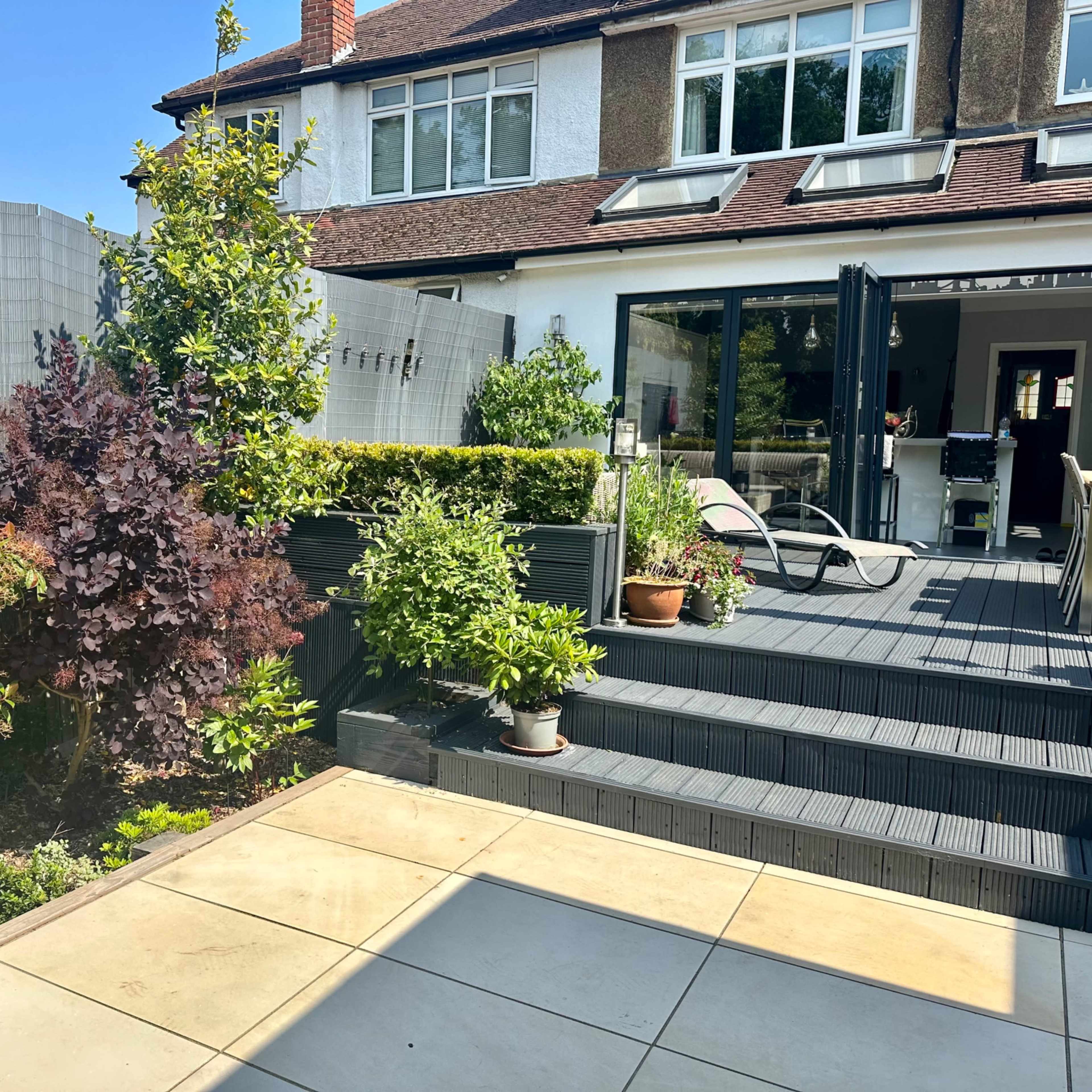 A landscaped backyard features a wooden deck with steps leading up to a modern home, surrounded by various plants and a privacy screen.