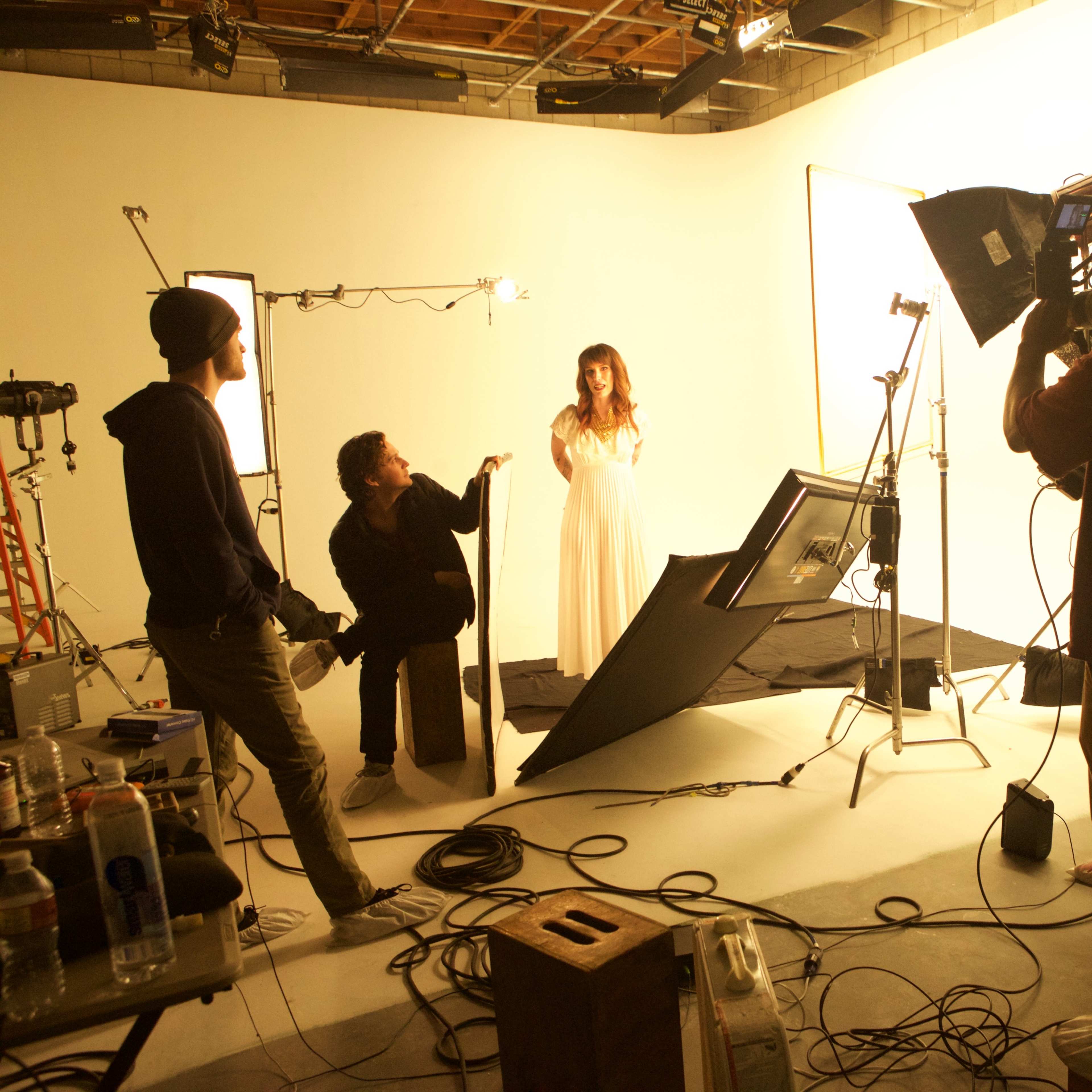 A film crew sets up a photoshoot in a studio with bright, white backdrops and various lighting equipment.