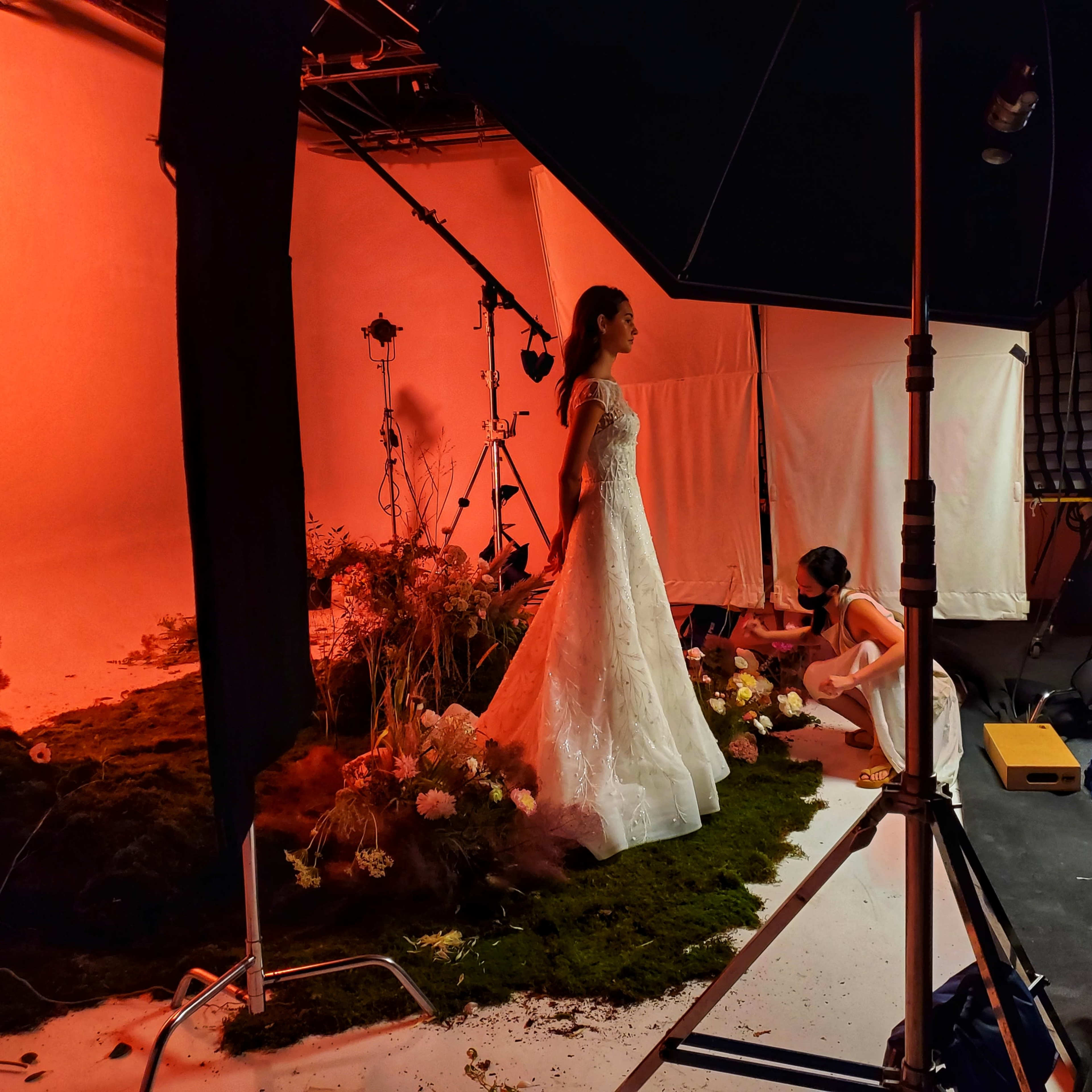 A model in a wedding dress stands in a studio surrounded by floral arrangements and lighting equipment.