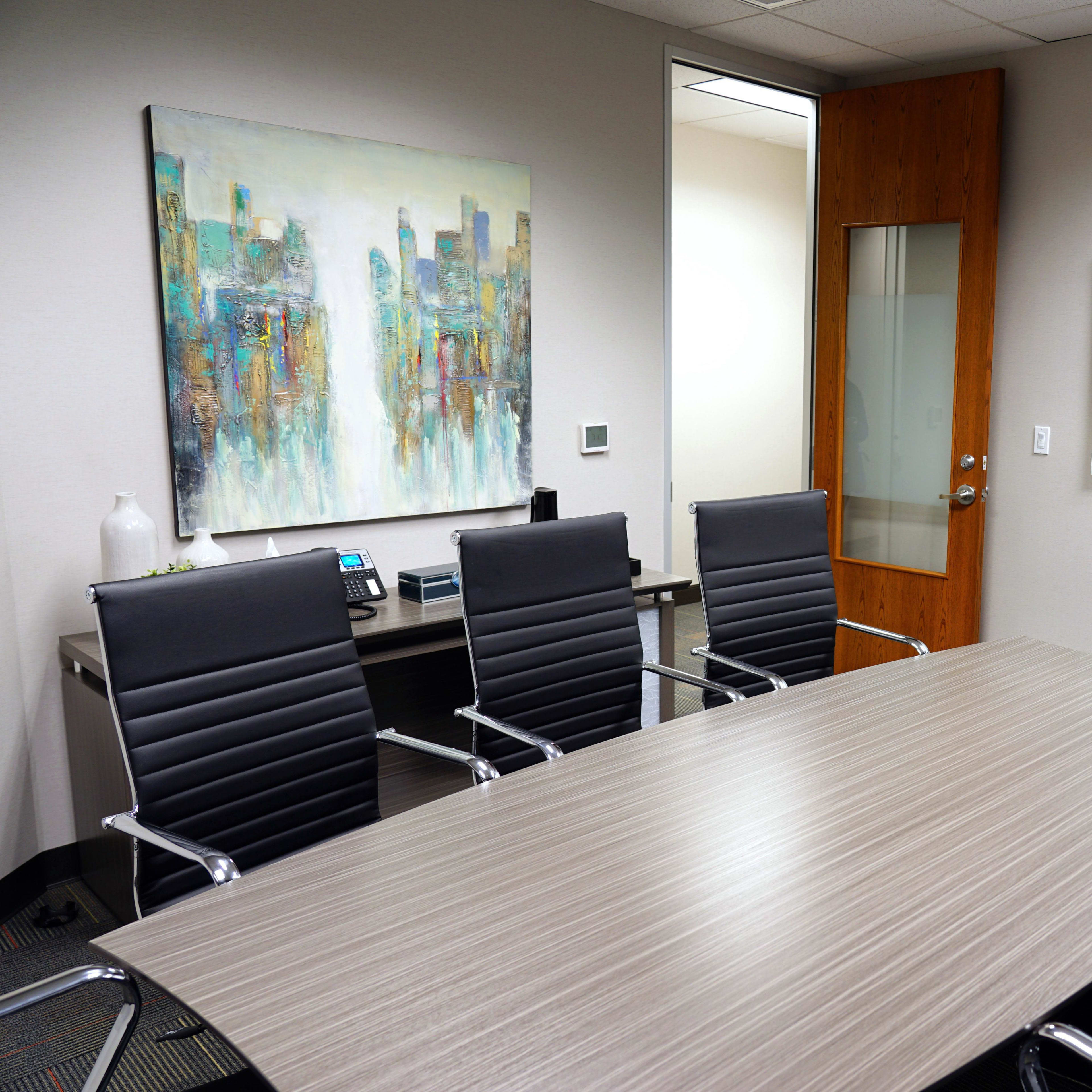 A modern conference room features a large oval table surrounded by eight black leather chairs, with a colorful abstract painting on the wall and a whiteboard nearby.