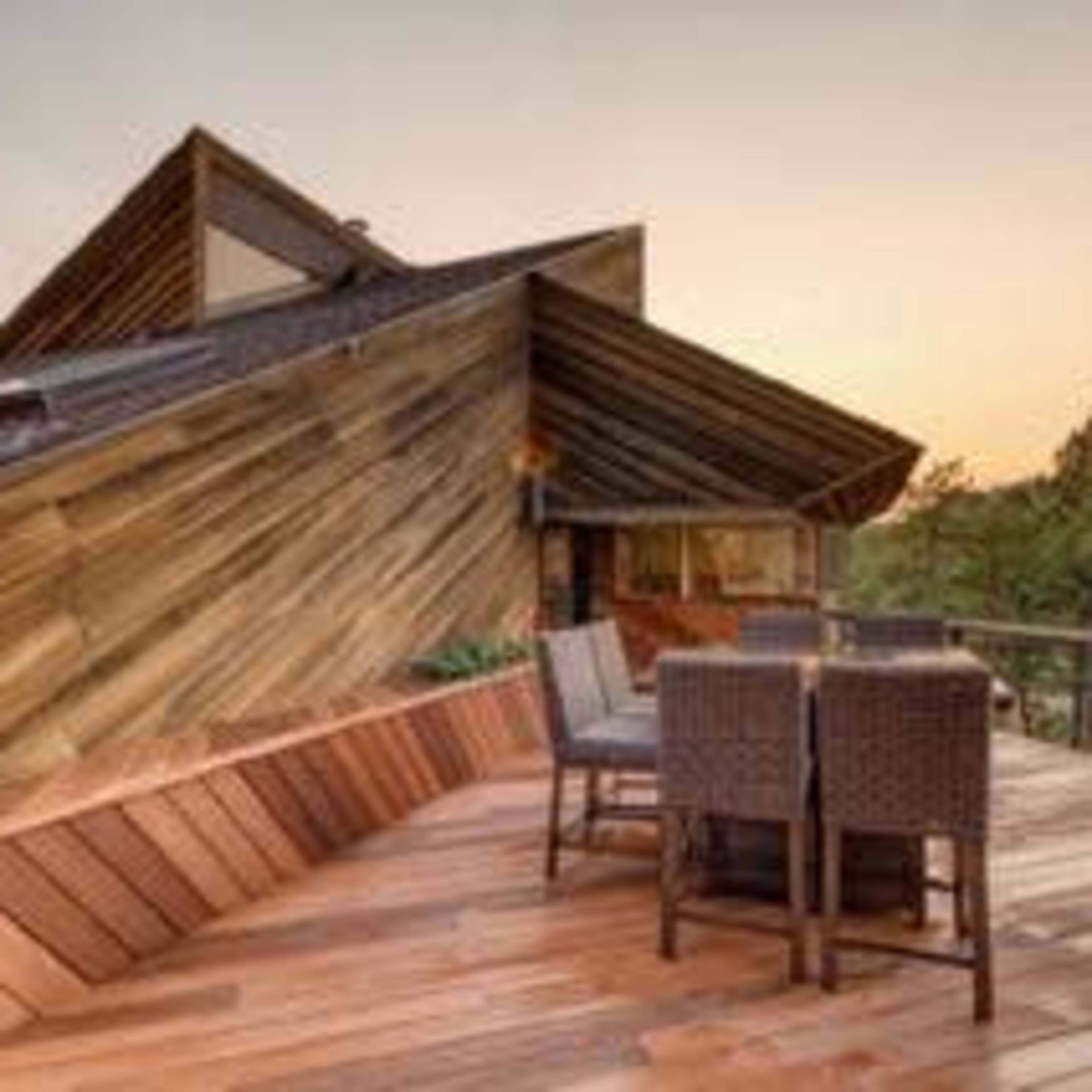 A wooden deck with a dining set is positioned next to a modern house featuring an angular roof design, surrounded by trees at sunset.