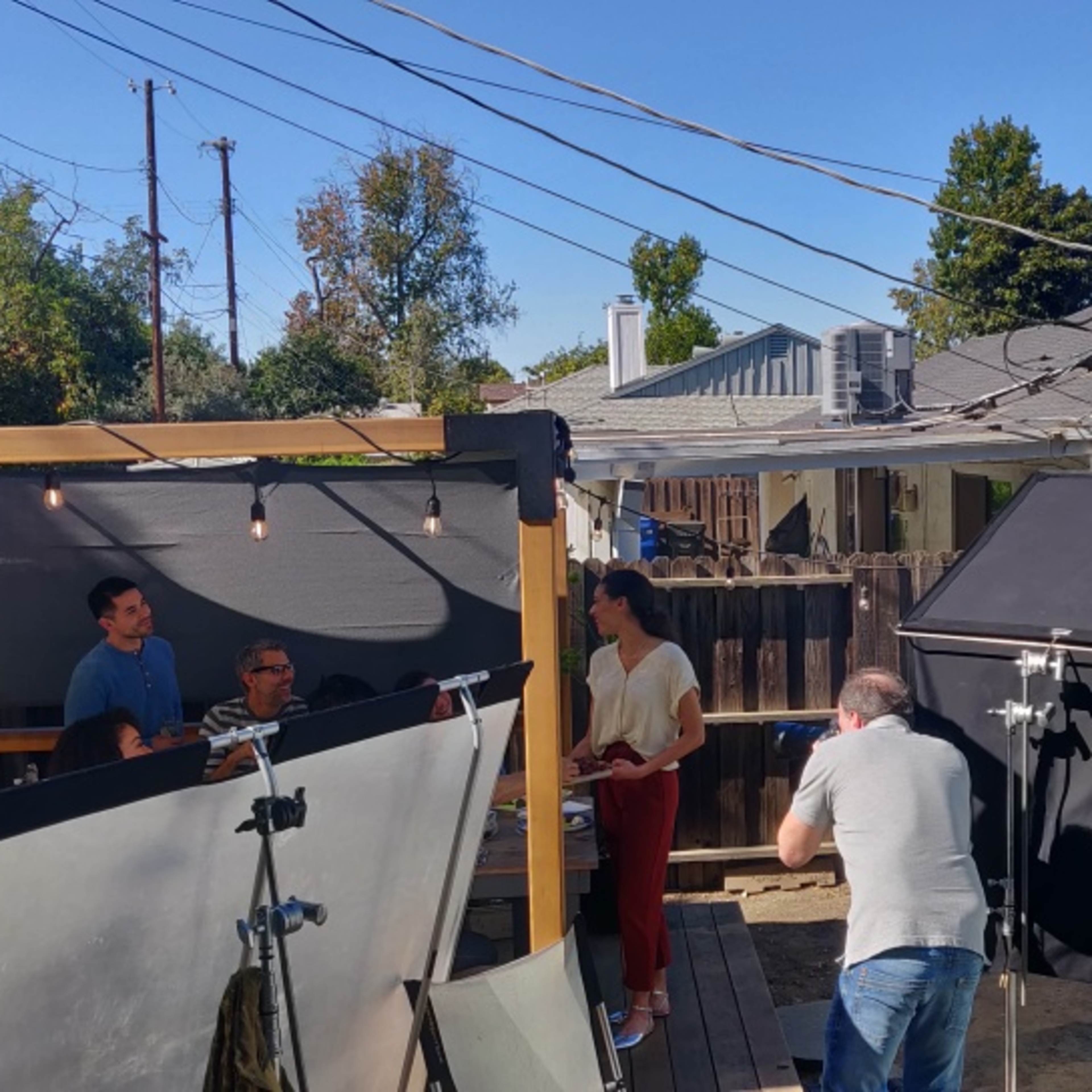 A film crew is set up in a backyard with lights and reflectors, filming a scene with several people engaged in conversation.