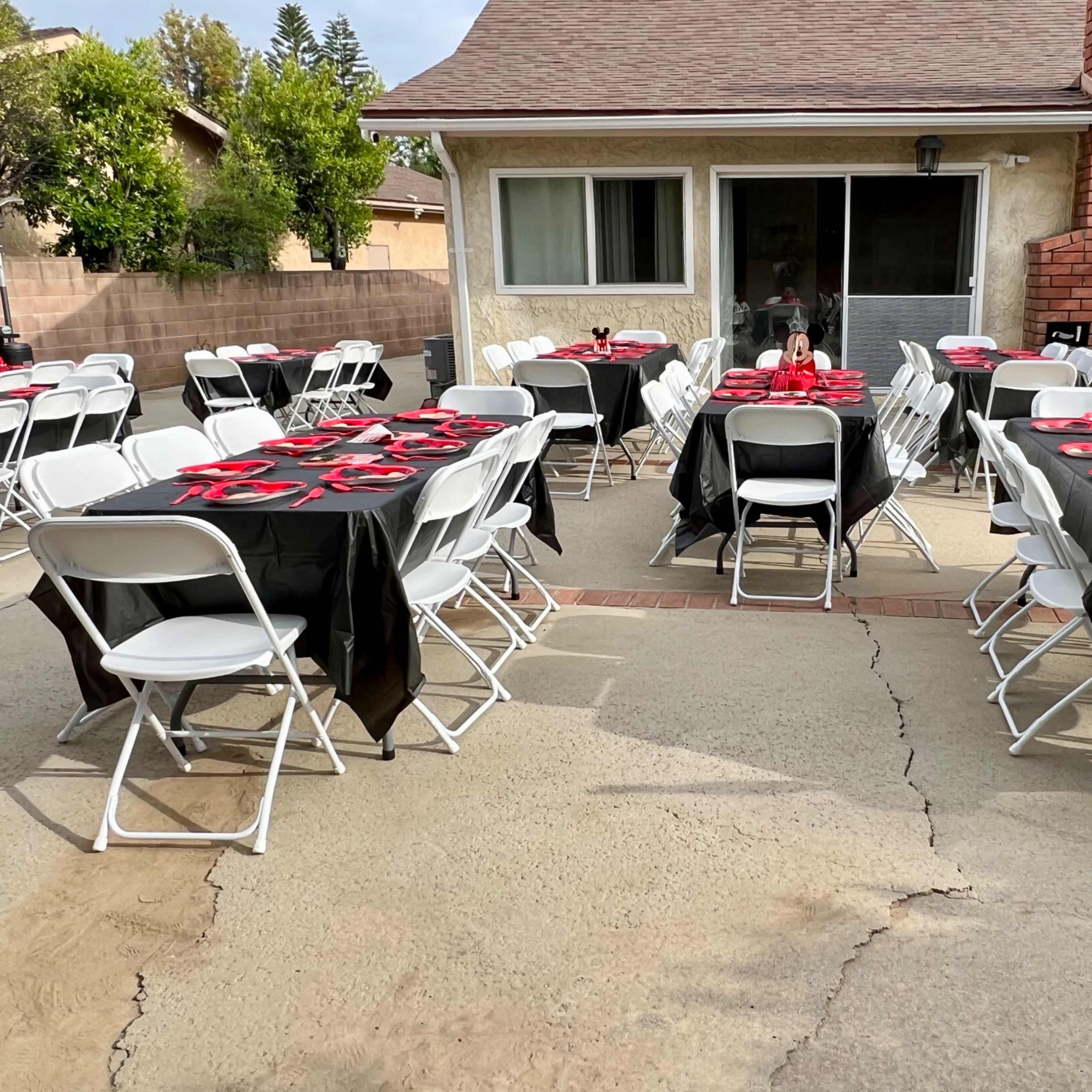 A backyard is set up with several tables covered in black tablecloths and red plates, surrounded by white folding chairs.