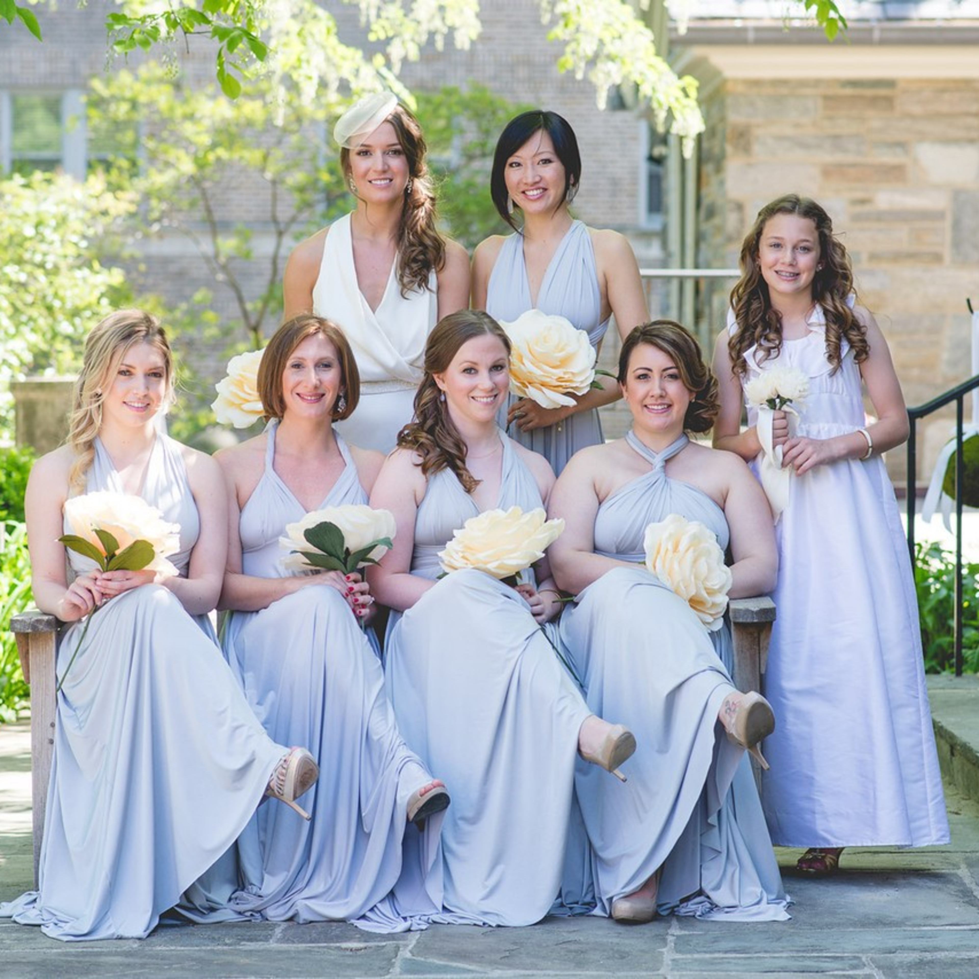 A group of seven women wearing light gray dresses poses for a photo in a garden setting.