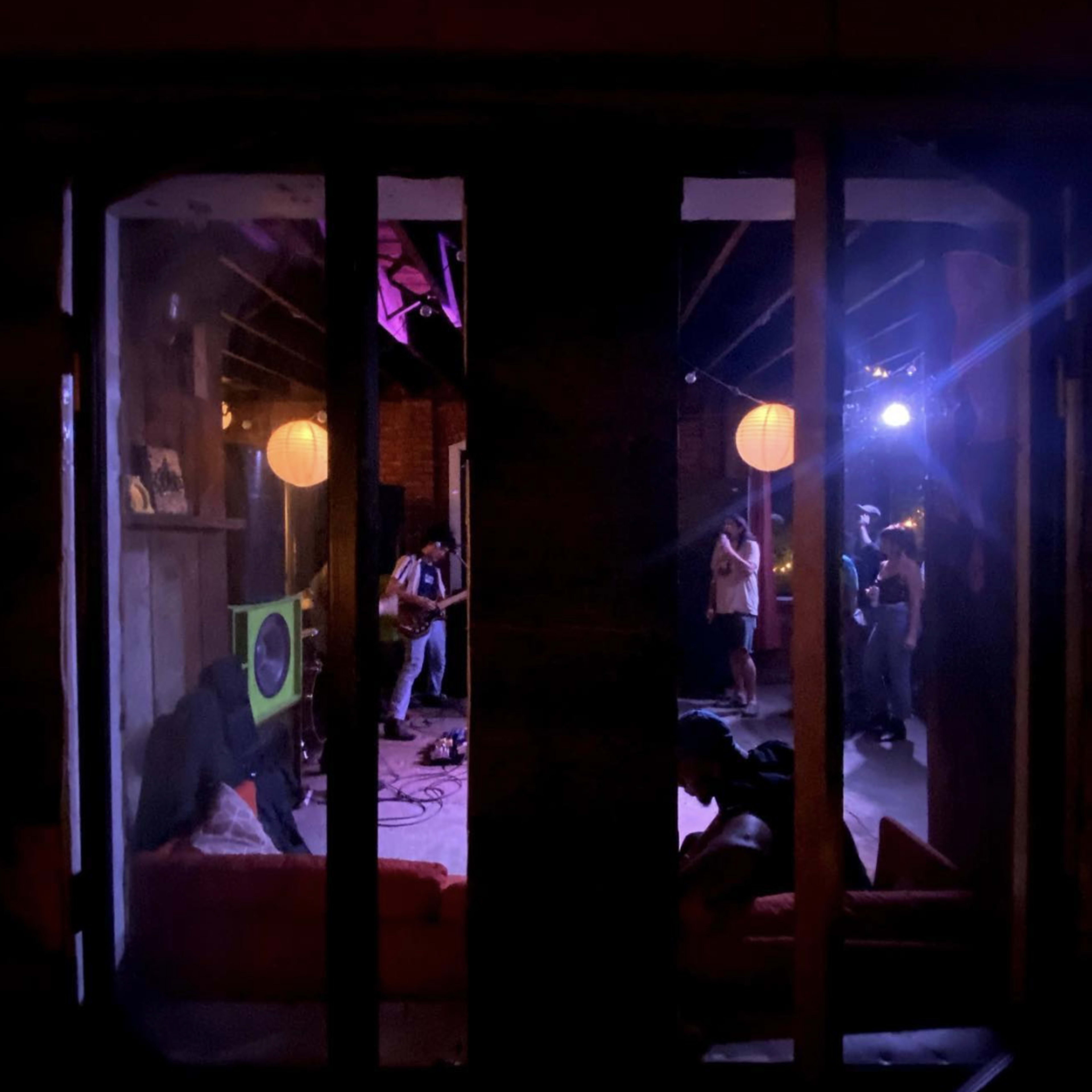 A dimly lit interior space with a band performing in the background while people socialize and relax on couches in the foreground.