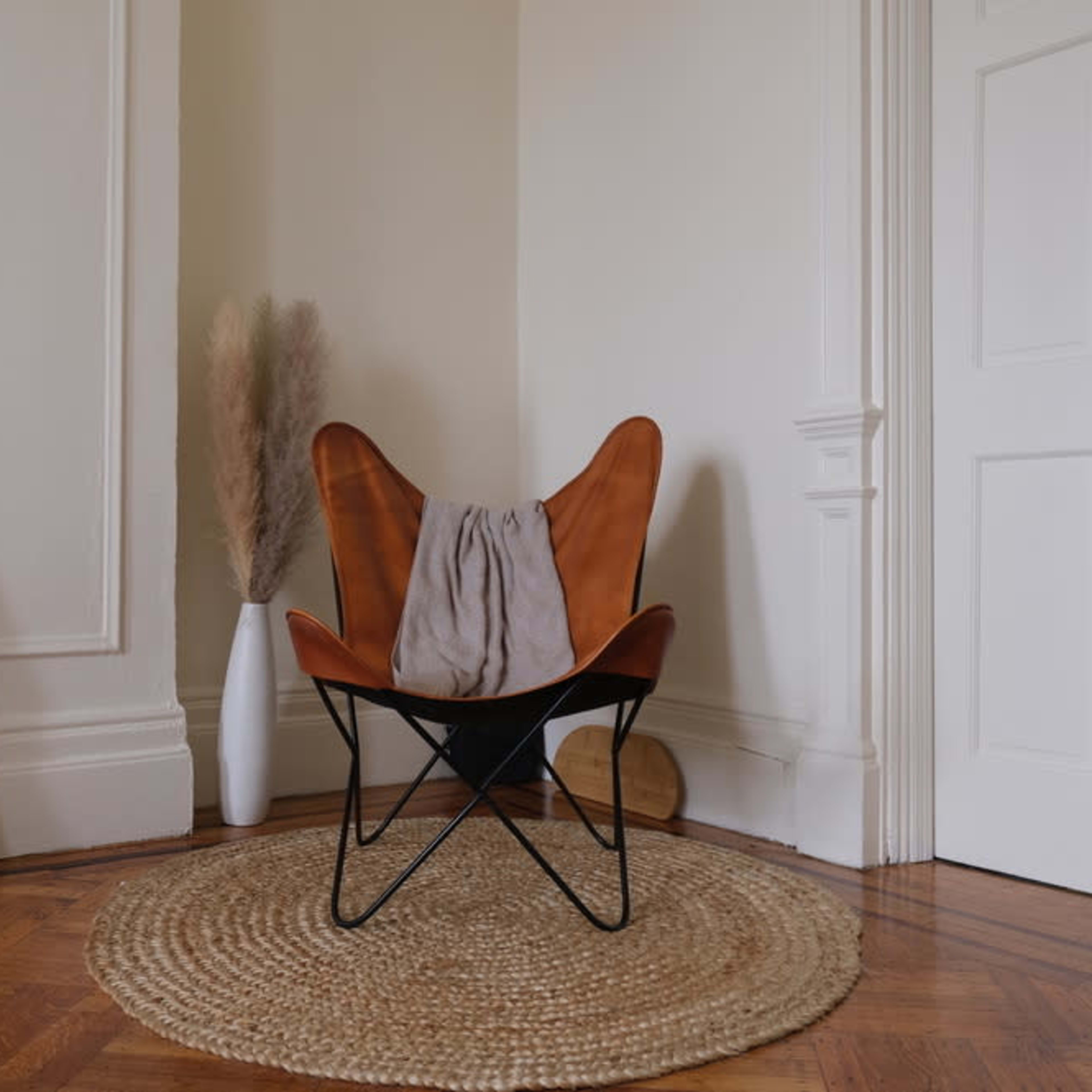 A butterfly chair with a gray draped fabric sits beside a framed line drawing and a tall vase with dried pampas grass on a circular rug in a room with light-colored walls.