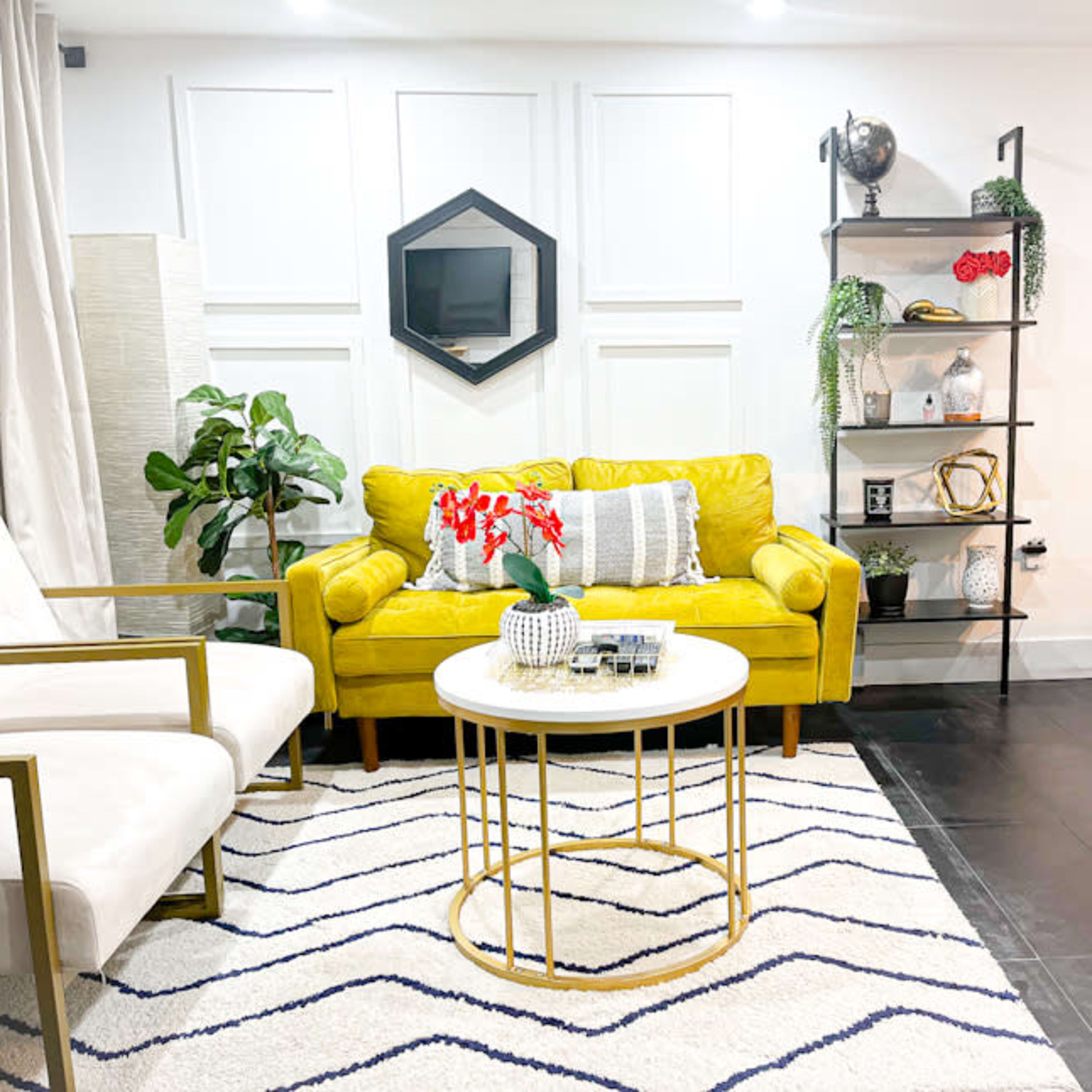 A modern living room with a yellow couch, white chairs, a round coffee table, and decorative shelving against a white wall.