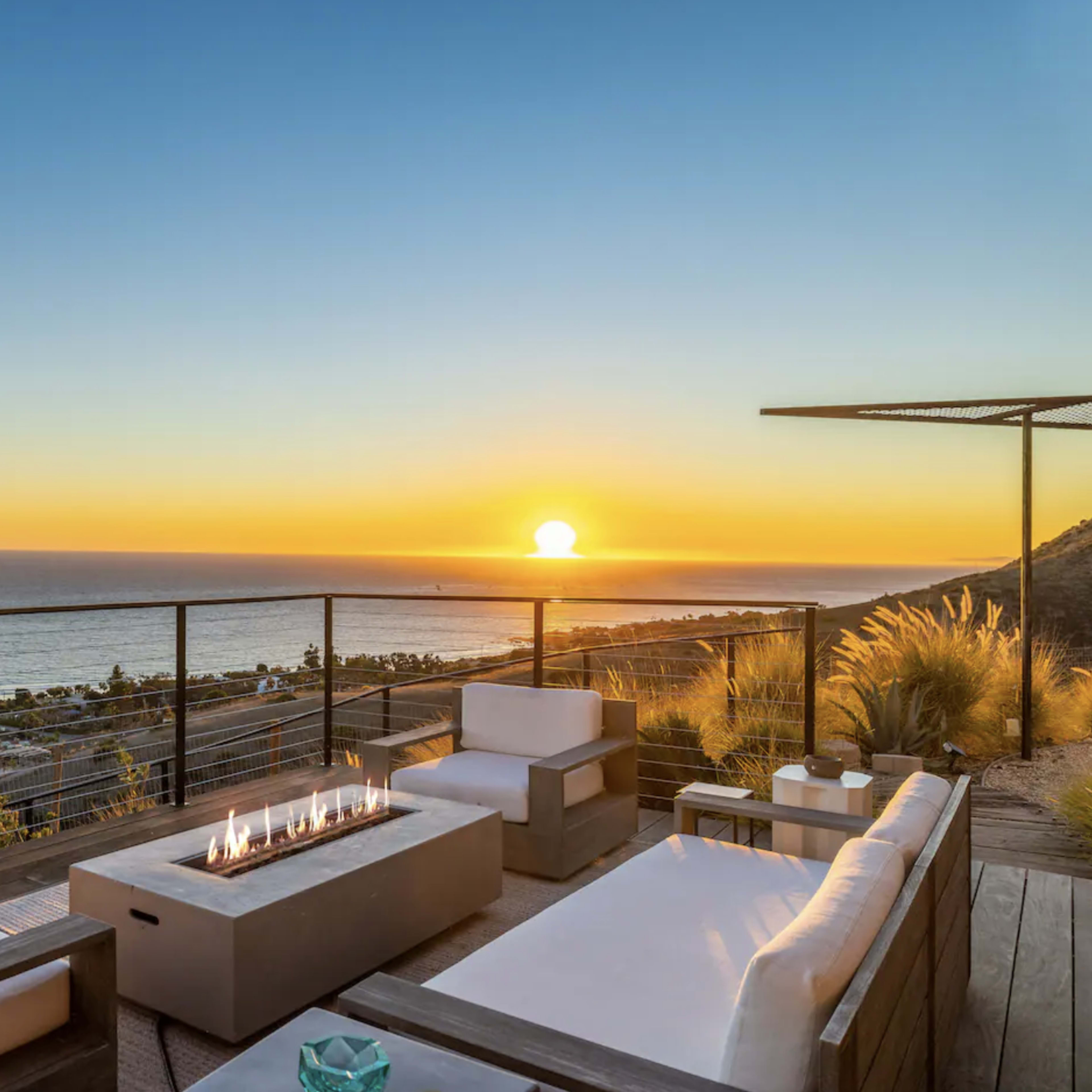 A modern outdoor seating area with a fire pit overlooks the ocean during sunset.