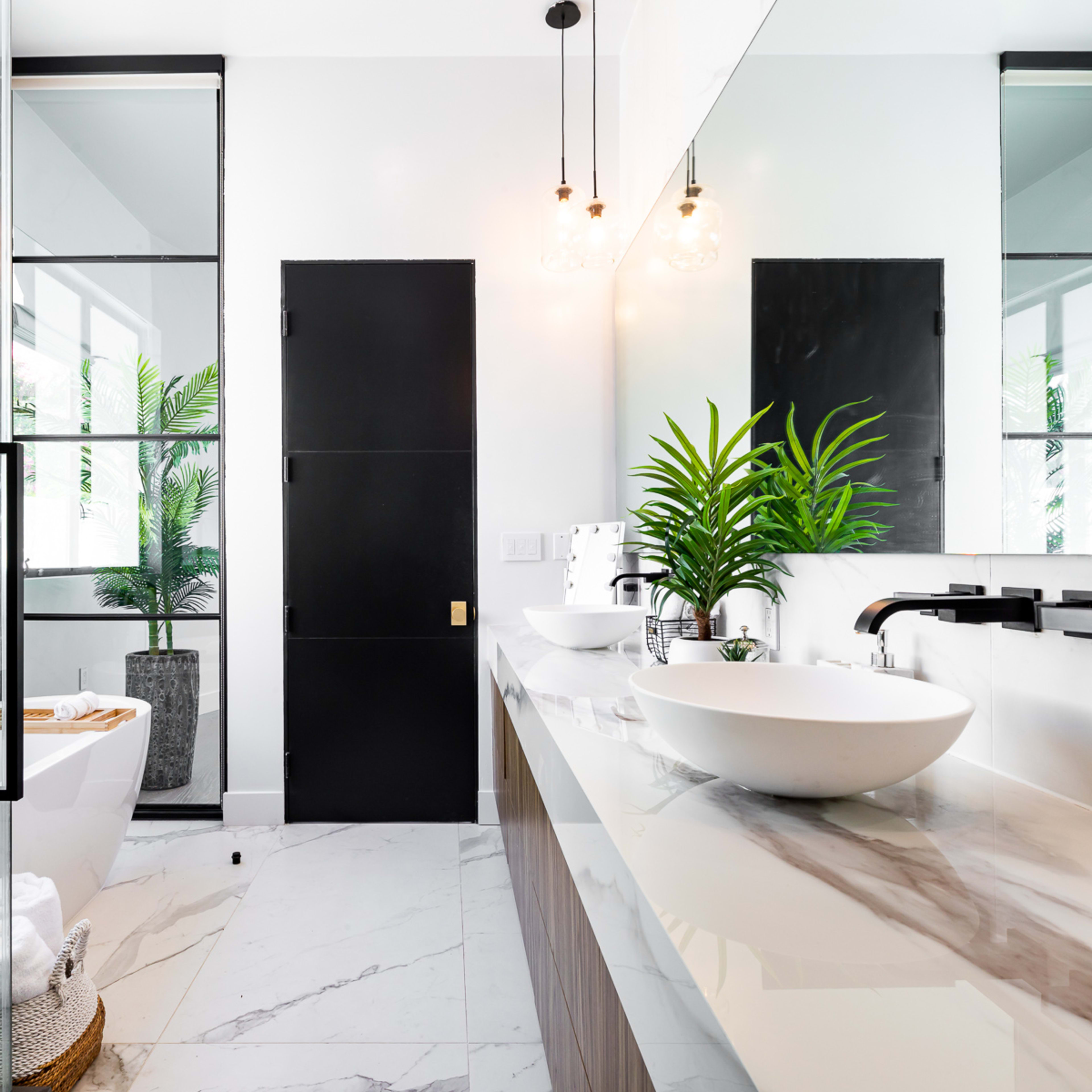 A modern bathroom featuring a freestanding bathtub, a large sink on a marble countertop, glass shower doors, and potted plants for decoration.