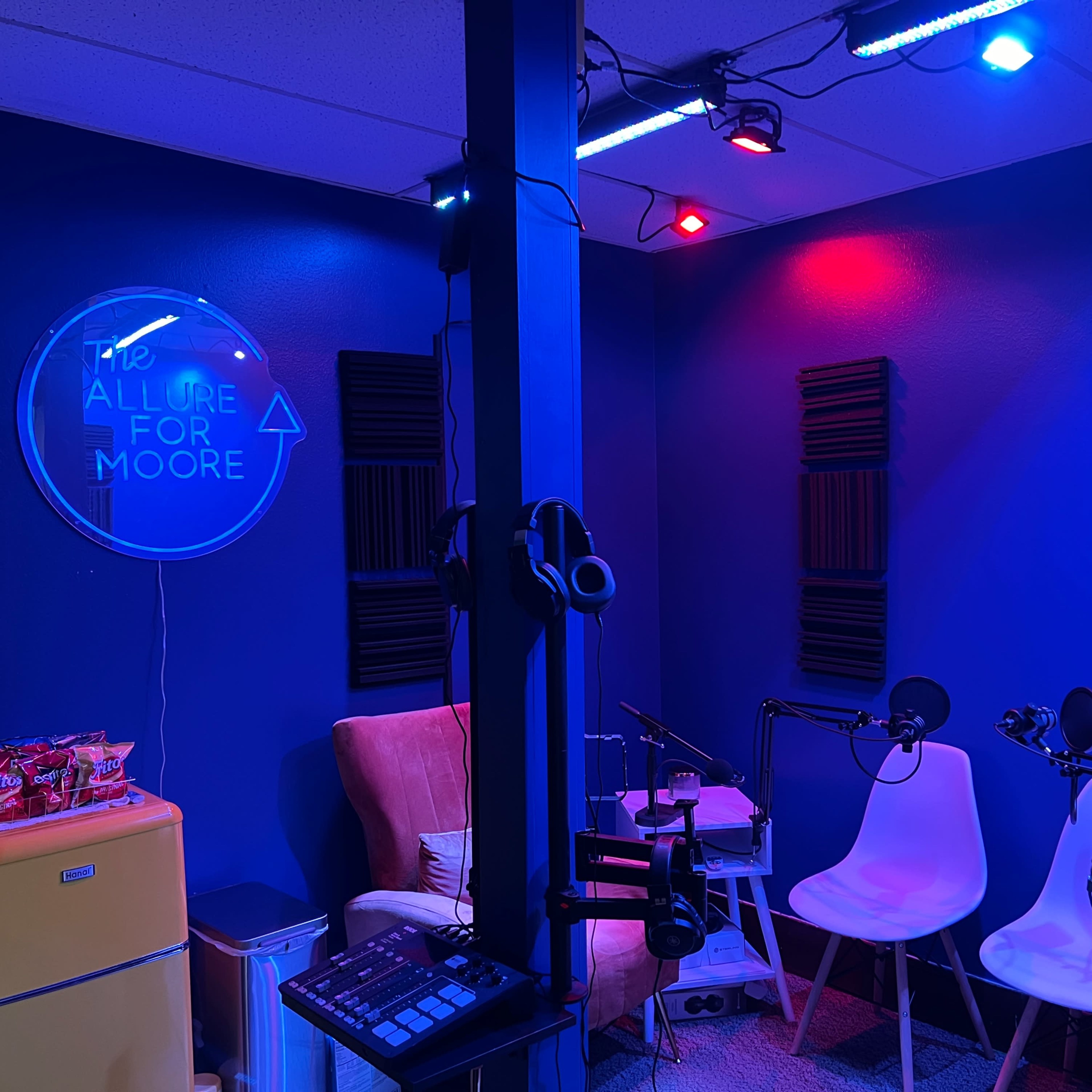A small recording studio features two microphones on stands, a cozy pink armchair, a mini fridge, and colorful lighting on the walls.