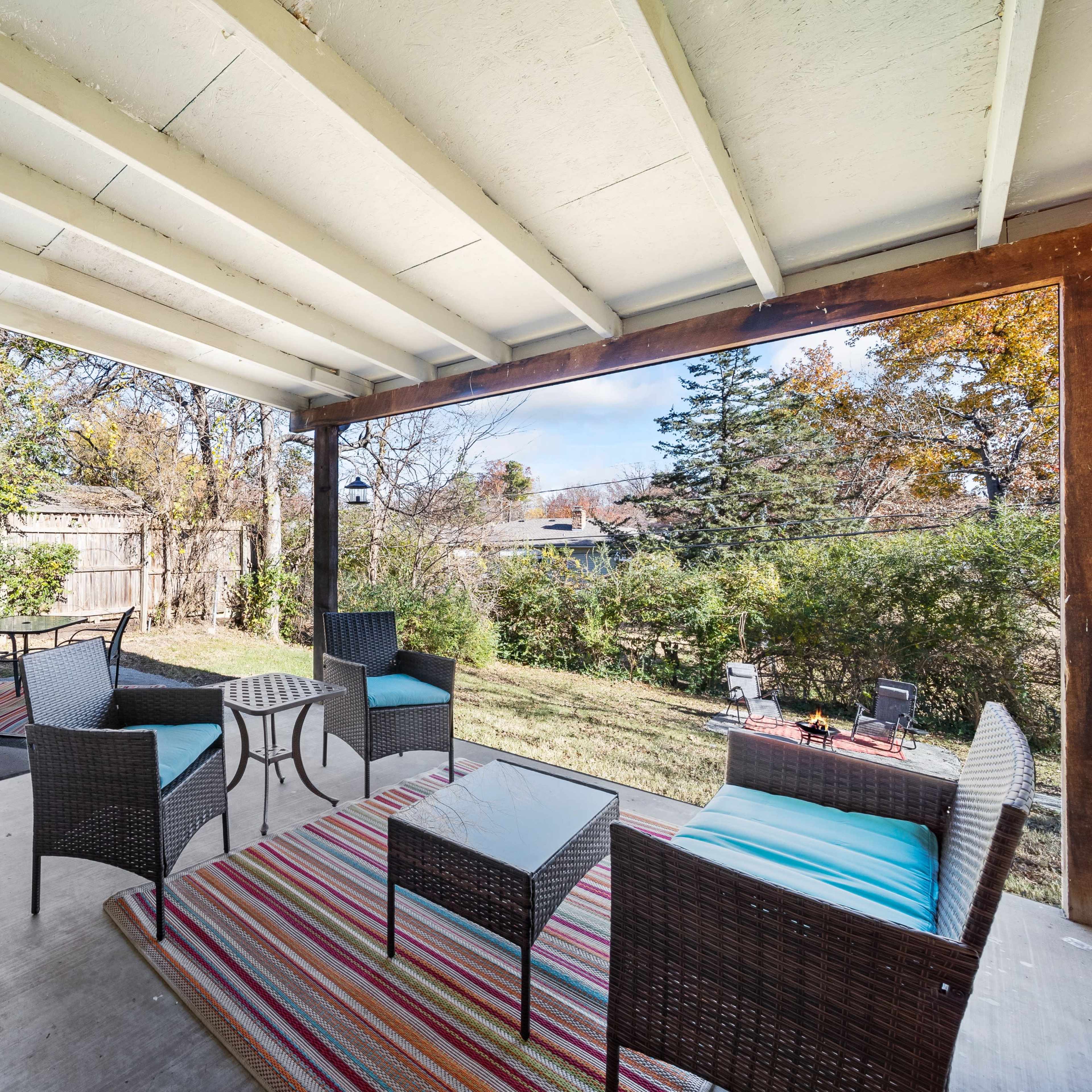 A covered patio features wicker furniture and a colorful rug, overlooking a backyard with trees and shrubs.