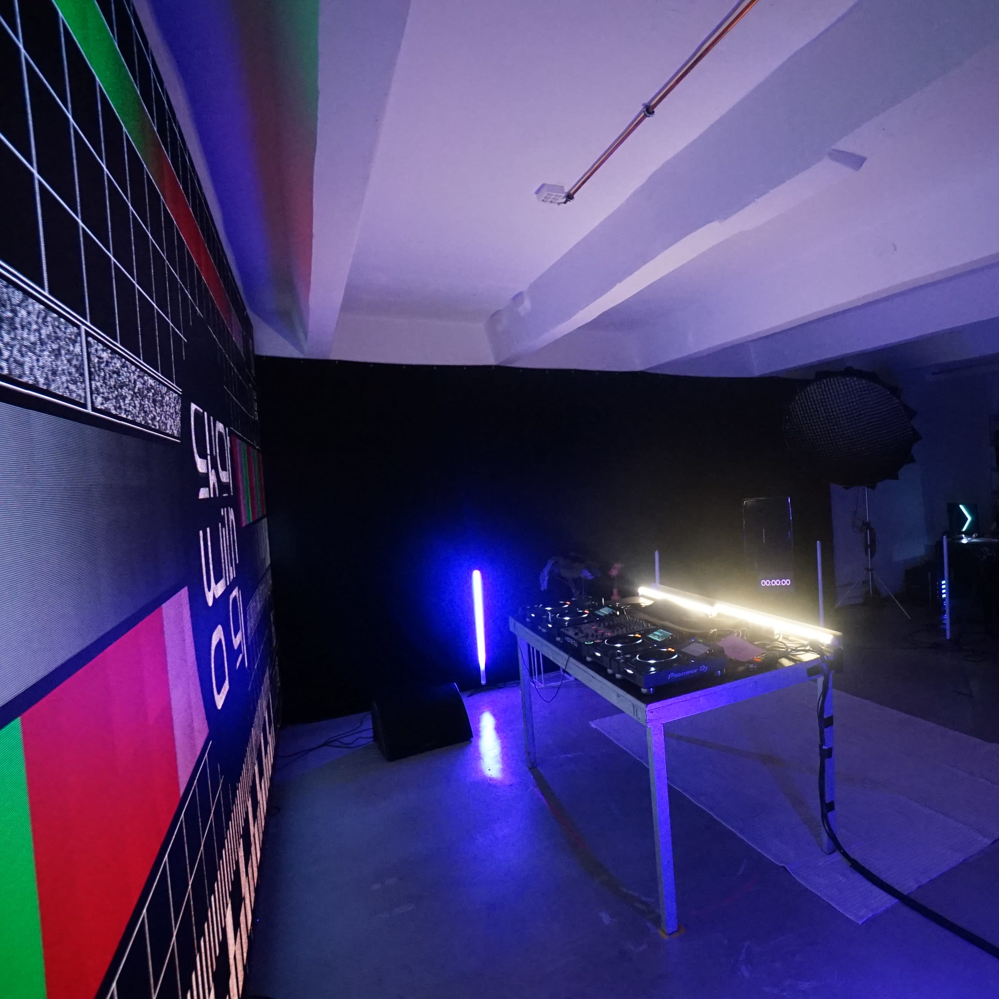 A DJ setup in a dimly lit room with a large screen displaying a test pattern and illuminated by colored lights.