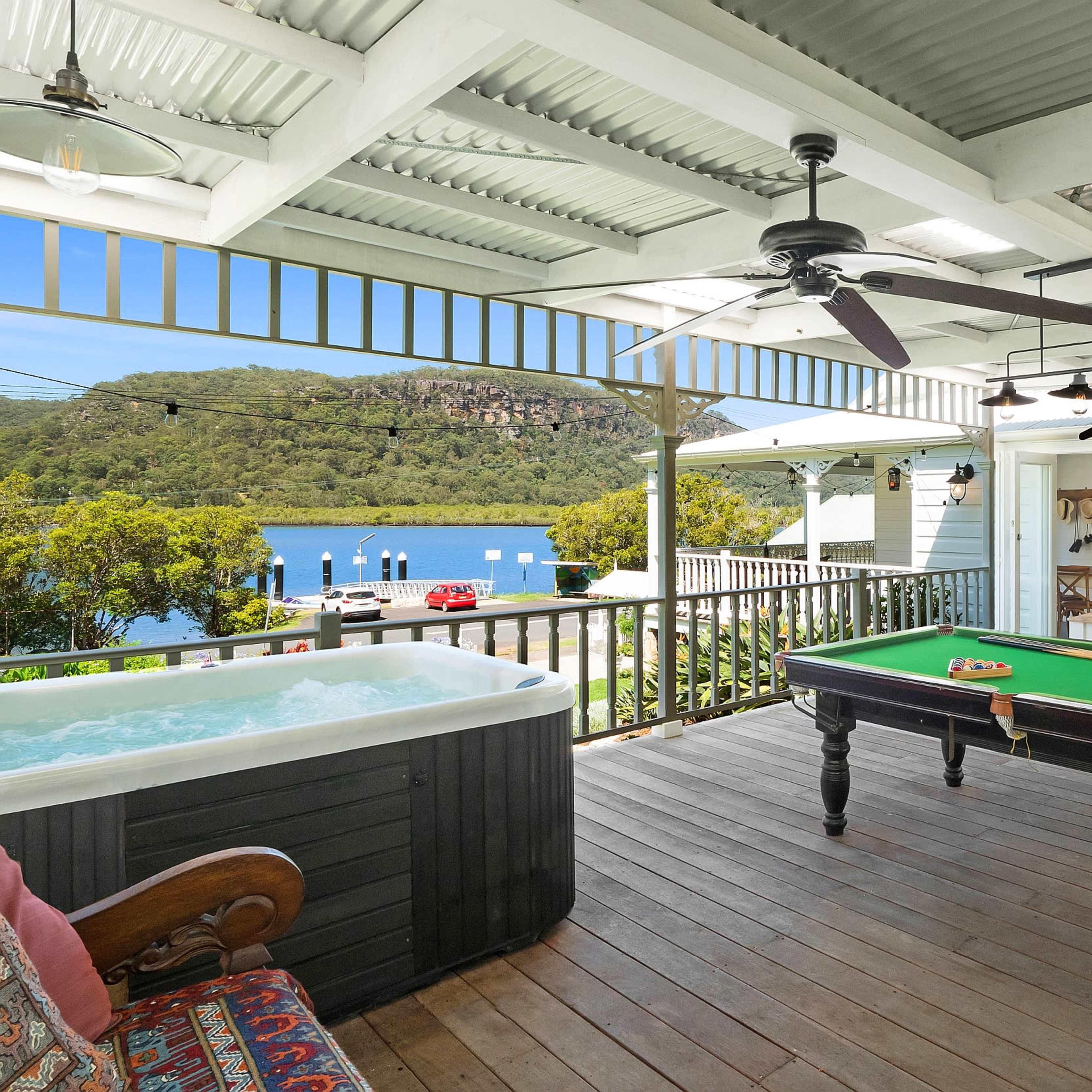 A spacious deck features a pool table and a hot tub, overlooking a waterfront with boats and lush greenery.