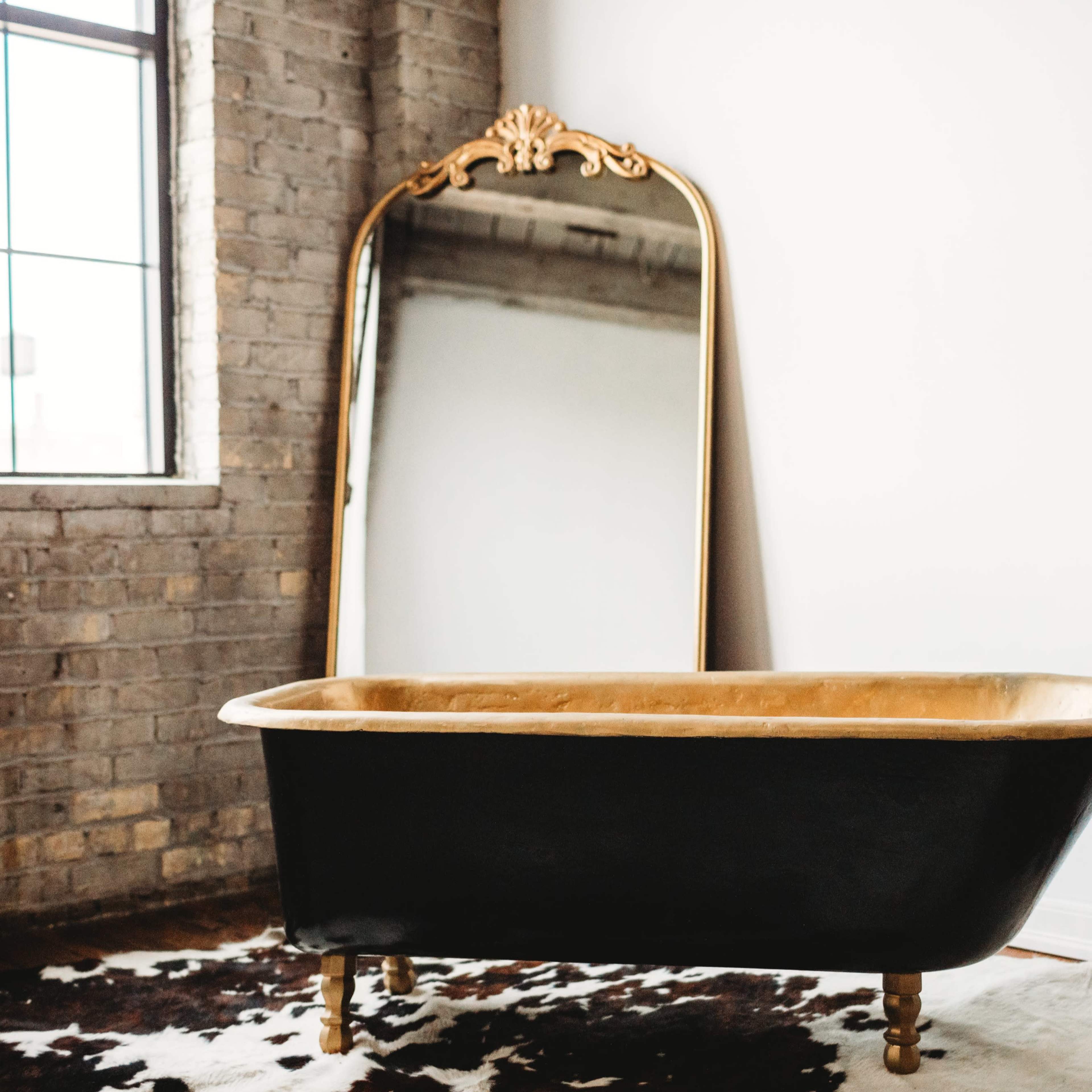 A black and gold freestanding bathtub is placed on a cowhide rug in front of a large, ornate mirror against a brick wall.