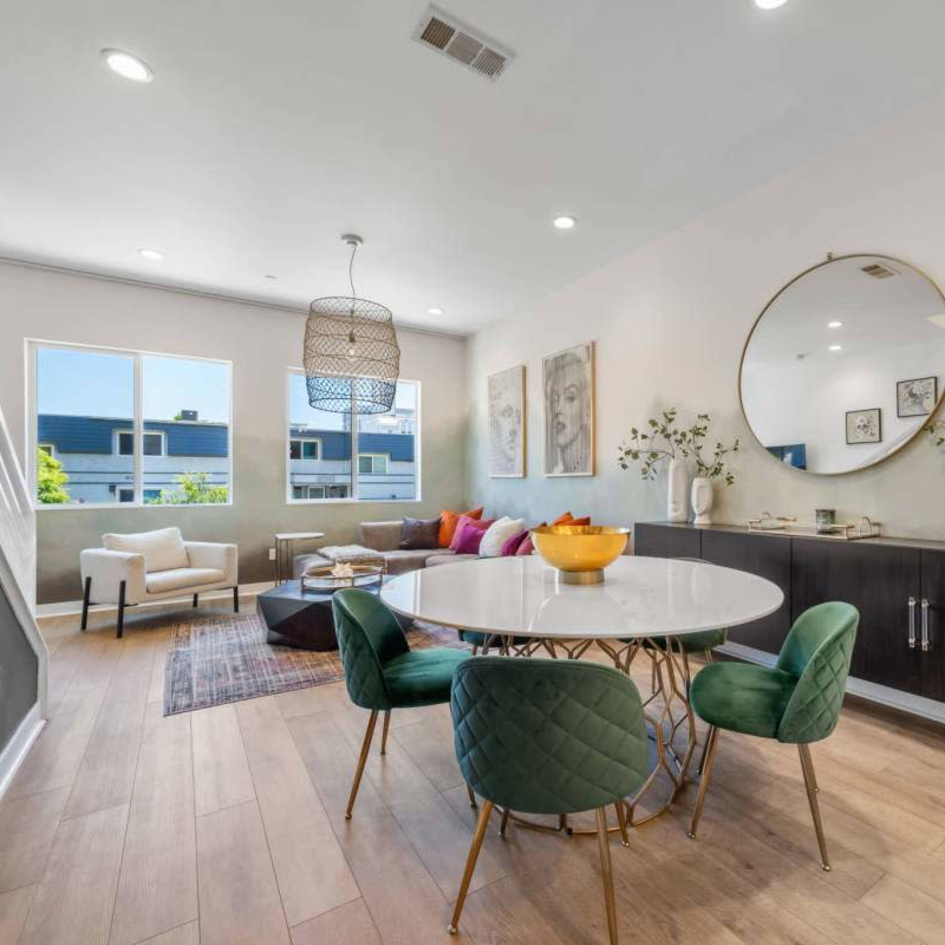 A modern dining area features a round table with green chairs, a large mirror, and a cozy seating arrangement in a well-lit space.