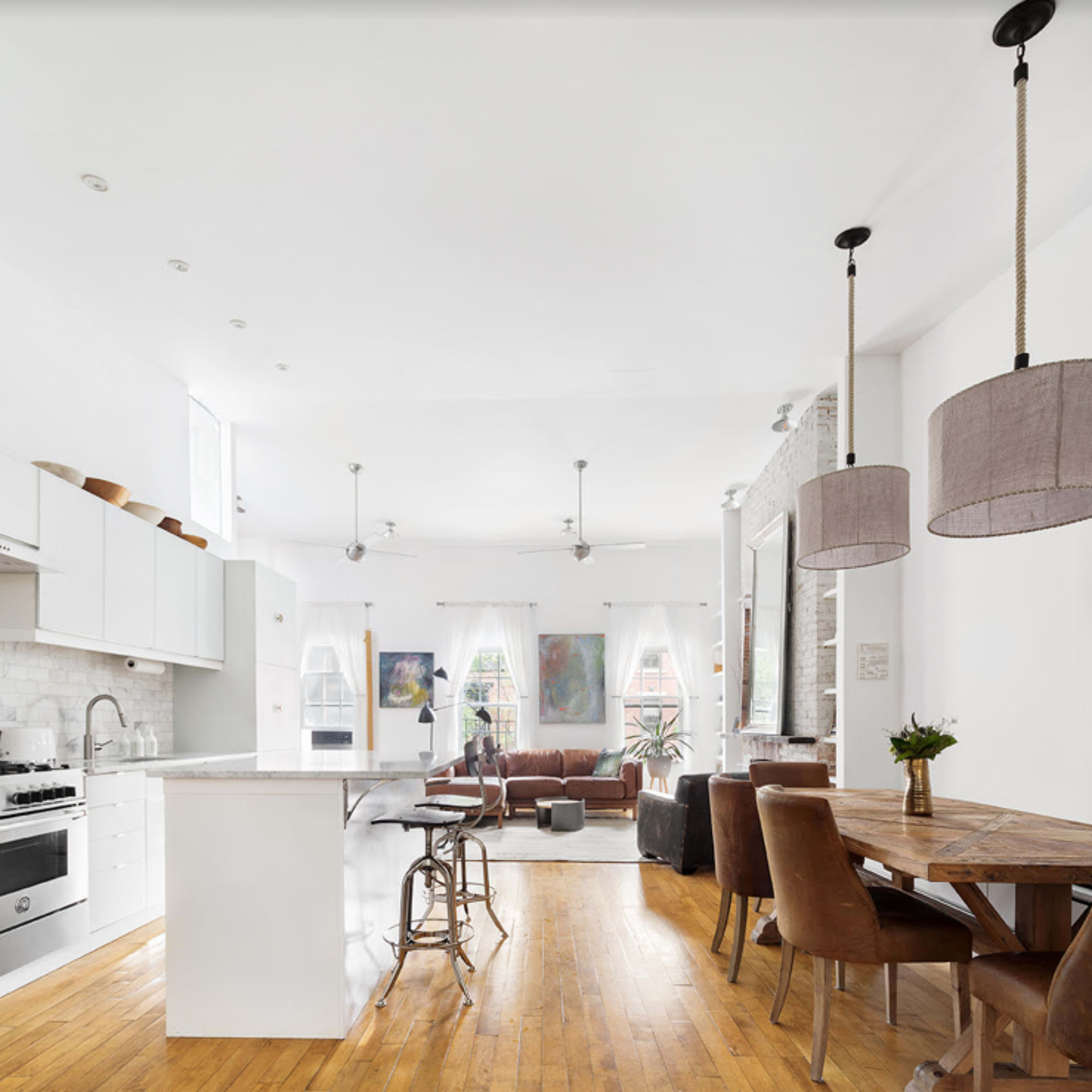 A modern kitchen and dining area with white cabinetry, a central island, a wooden dining table, and a brown leather sofa.