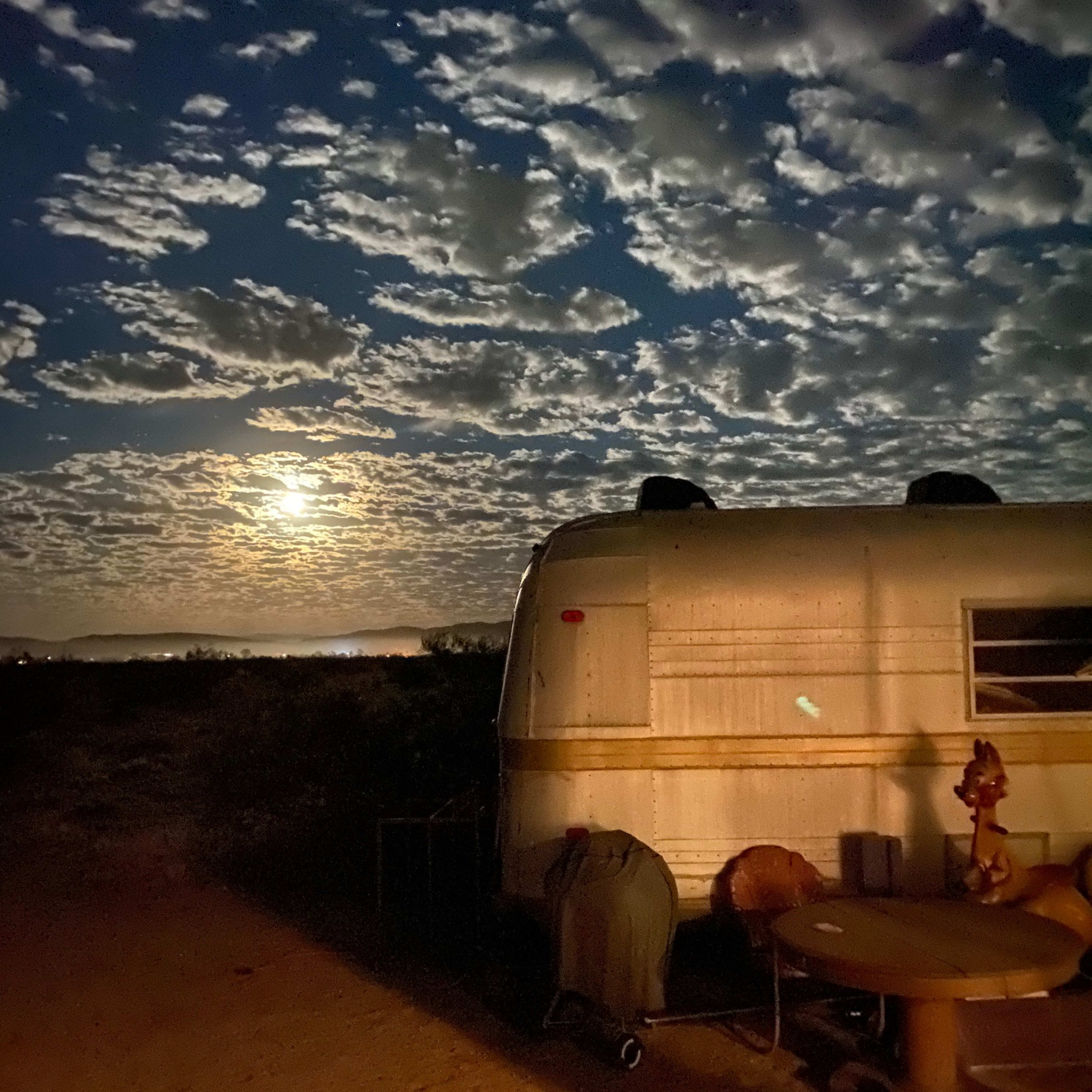 A vintage trailer is parked in a desert setting under a moonlit sky filled with clouds.
