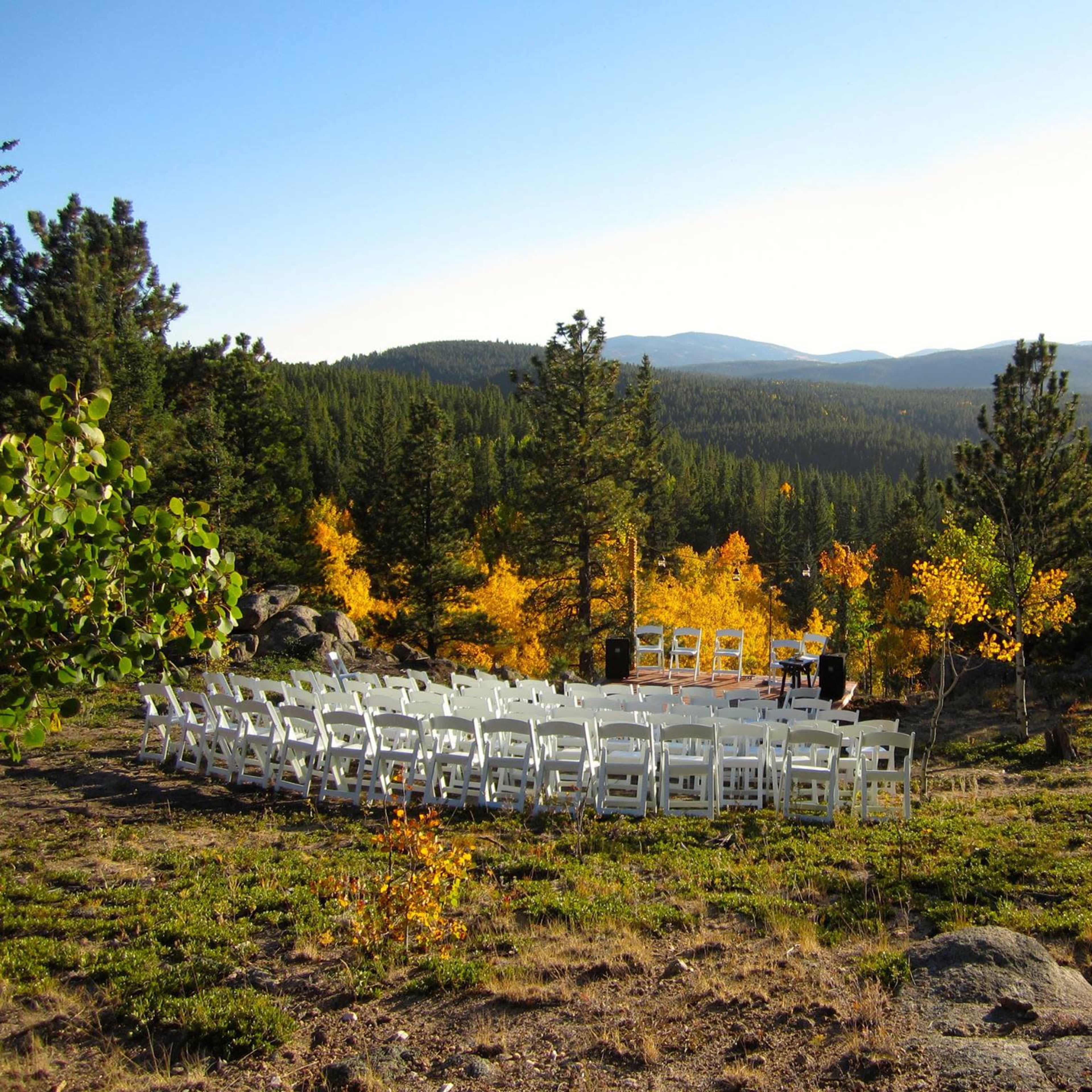 A circular arrangement of white chairs is set up on a grassy area surrounded by trees and mountains.
