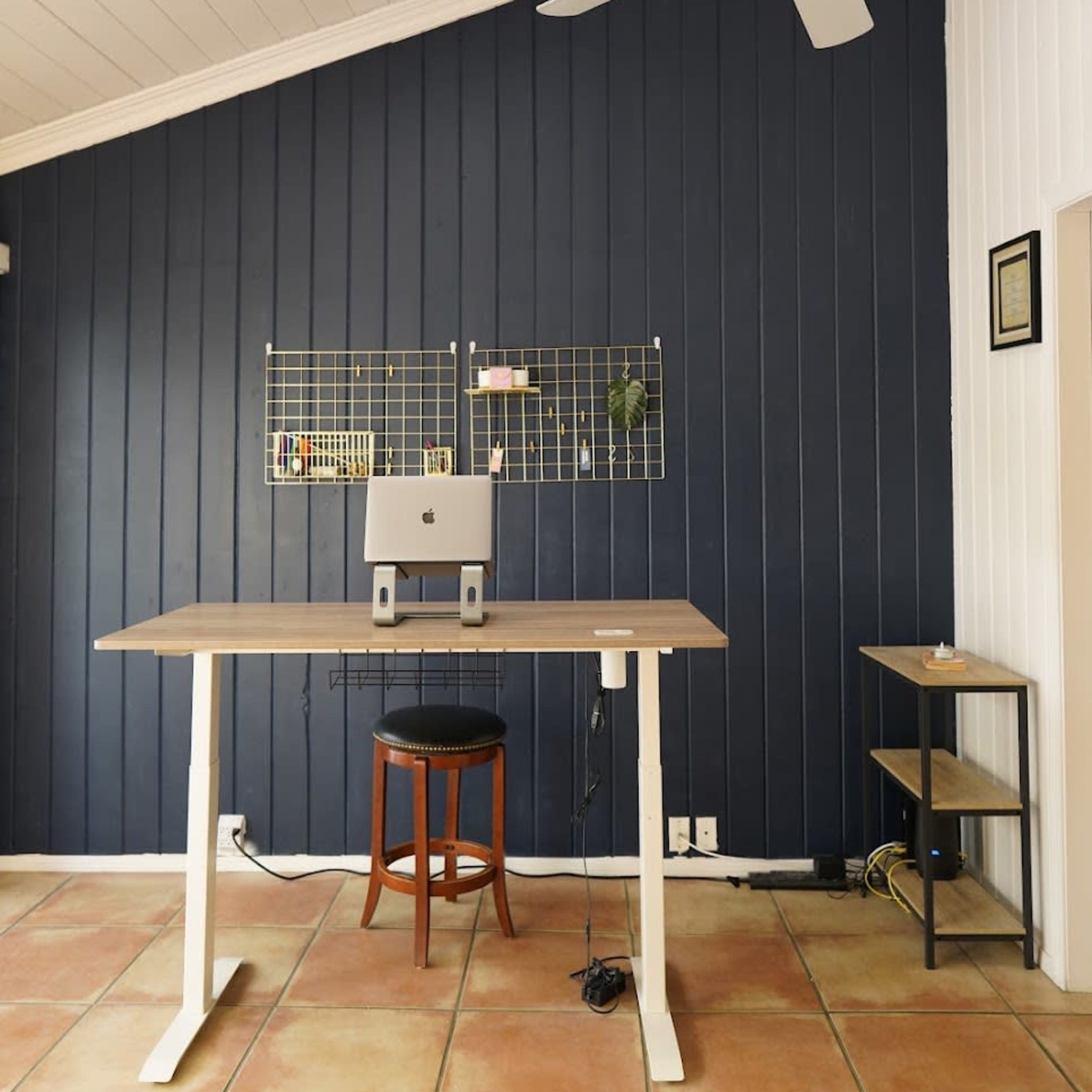 A minimalist workspace with a standing desk, a computer monitor, and a chair, set against a dark blue wall with white paneling and a ceiling fan above.