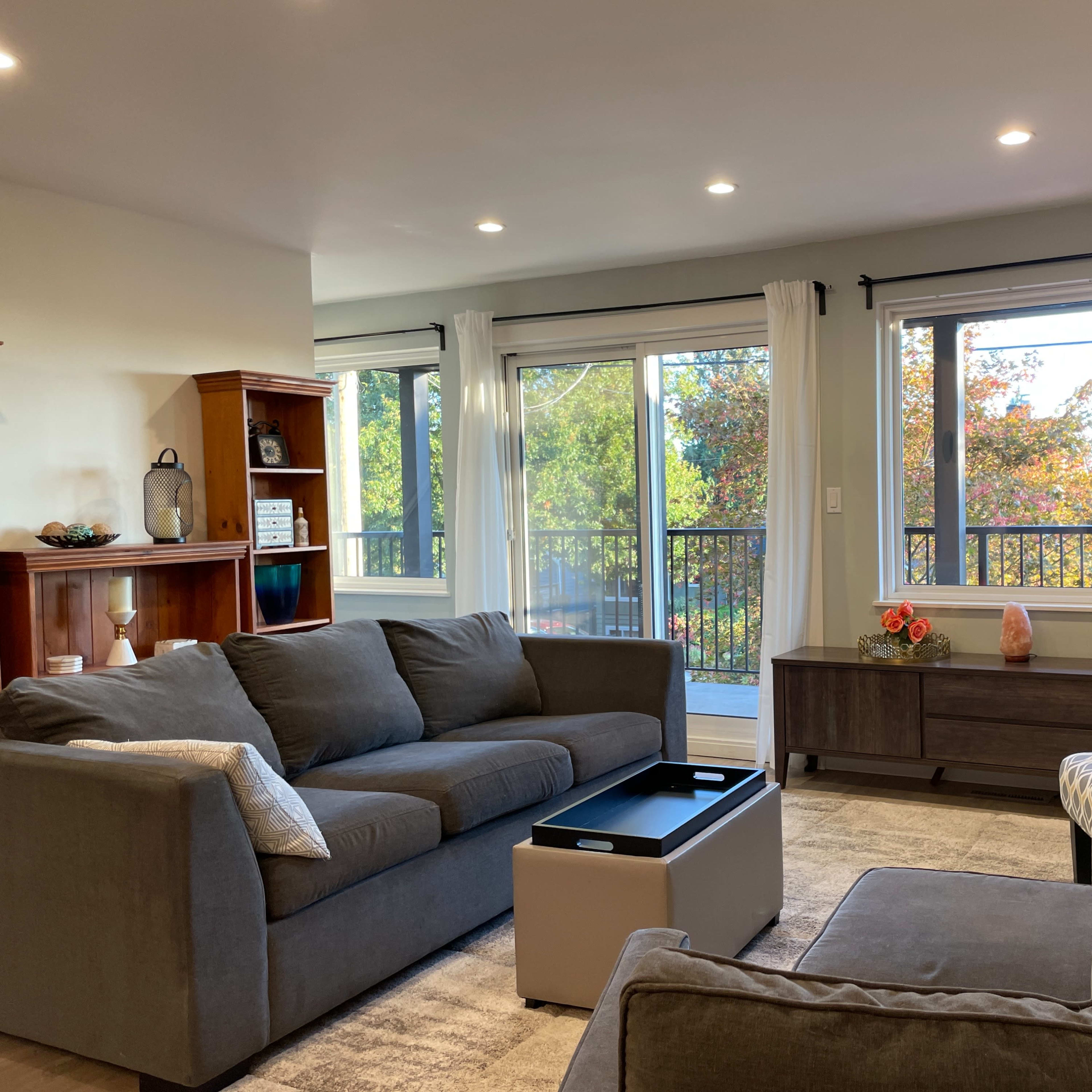 The living room features two gray couches, a coffee table, a mounted TV, and large windows overlooking a tree-filled view.