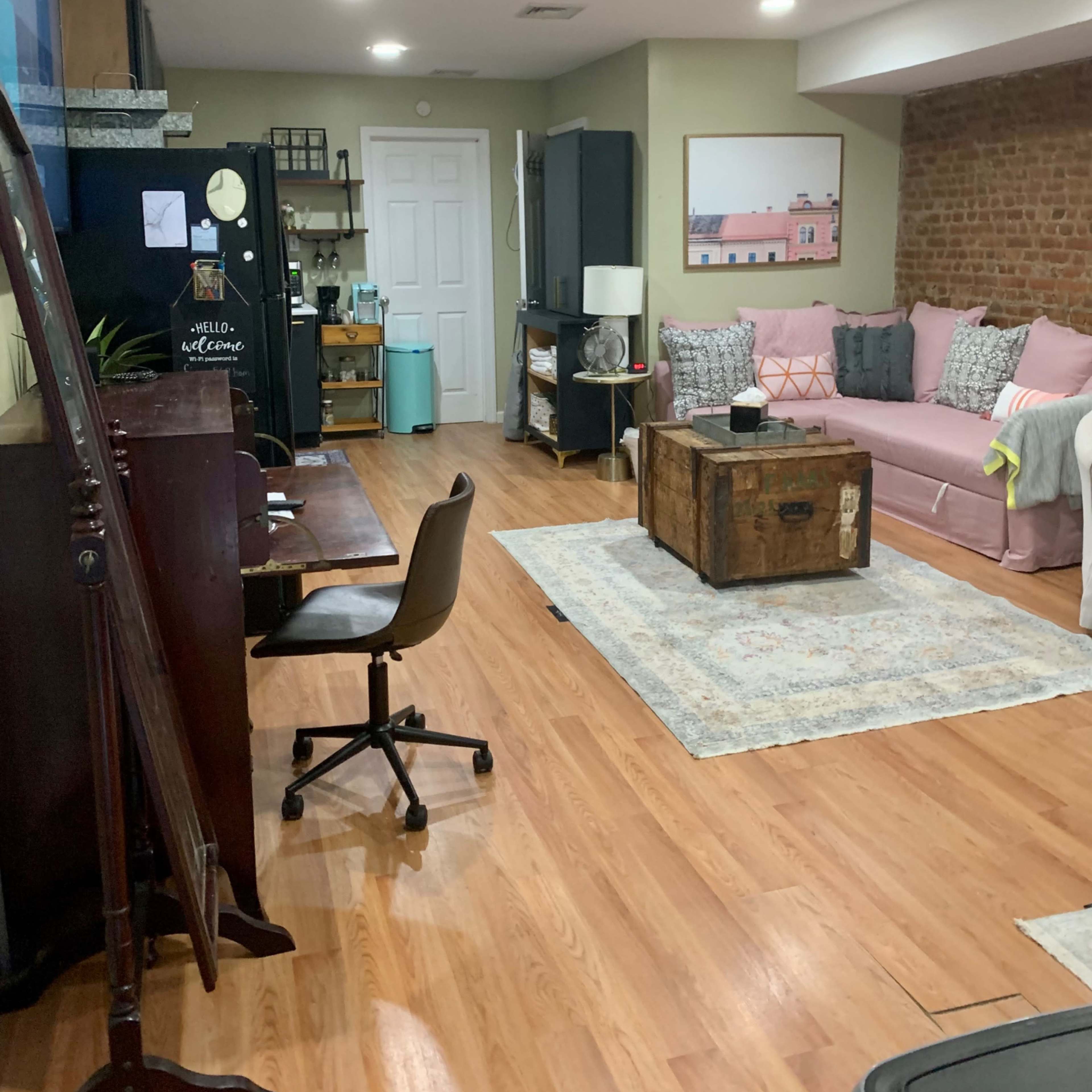A cozy living space with a pink couch, wooden coffee table, and a desk, along with a brick wall and various decorative items.