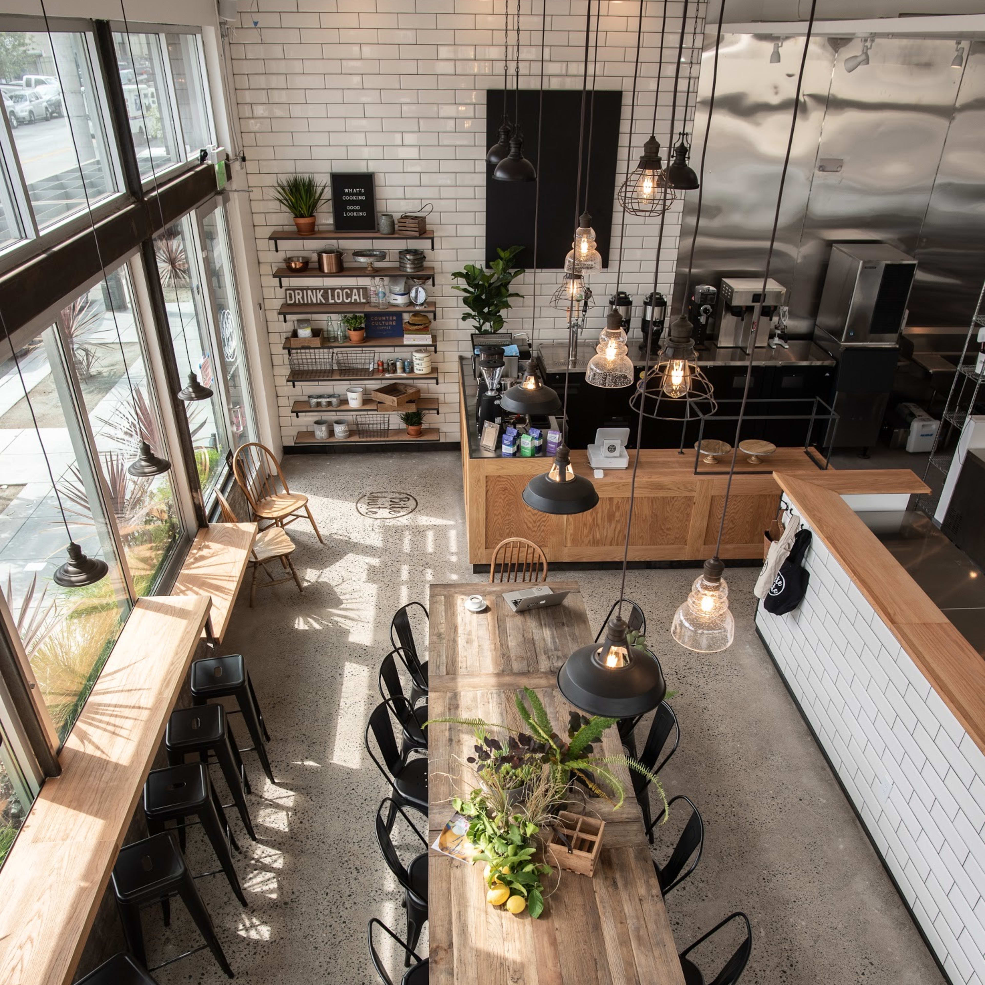 An industrial-style cafe with a long wooden table, black chairs, large windows, and pendant lighting.