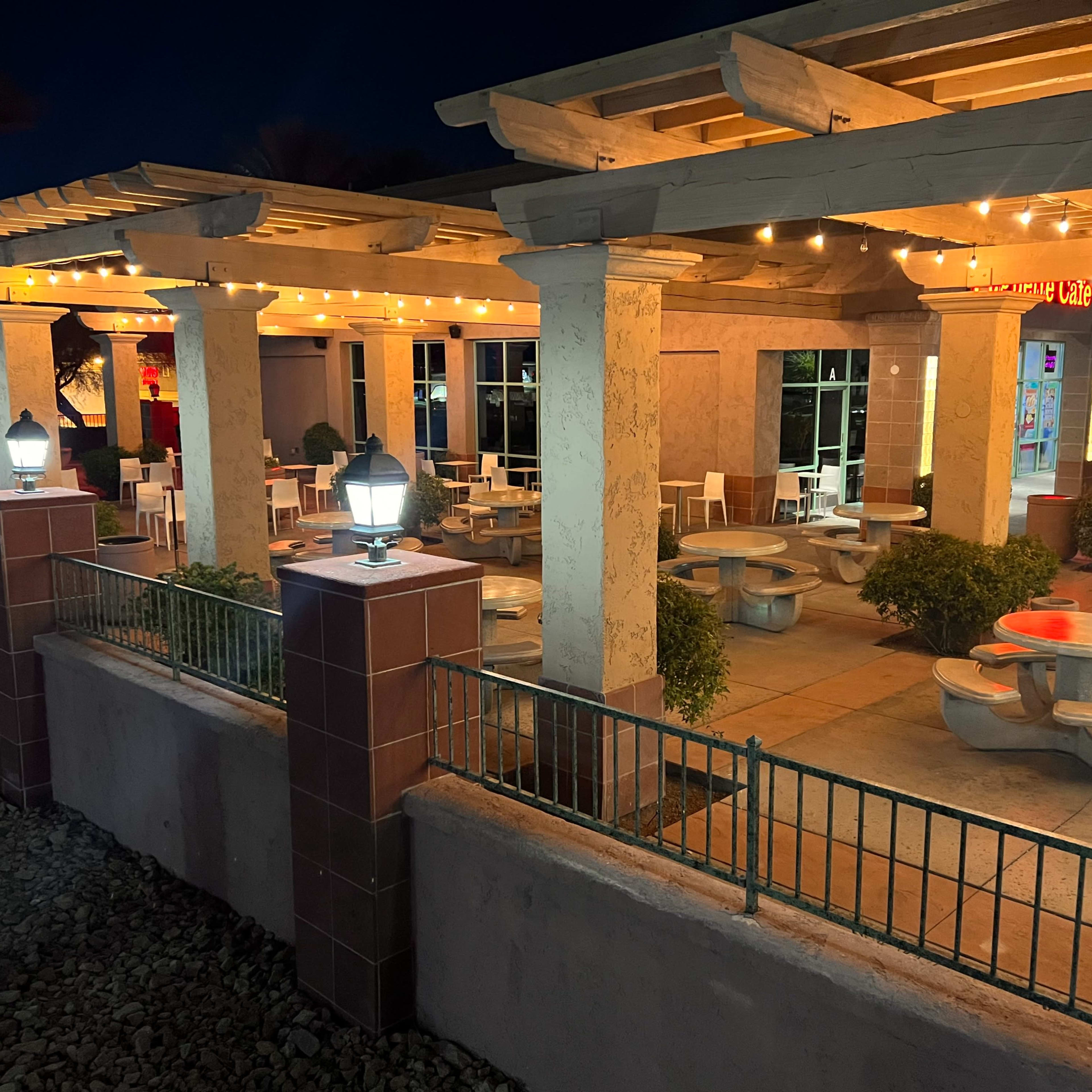 A partially illuminated outdoor dining area with several table and chair setups under a covered structure, framed by decorative lighting.