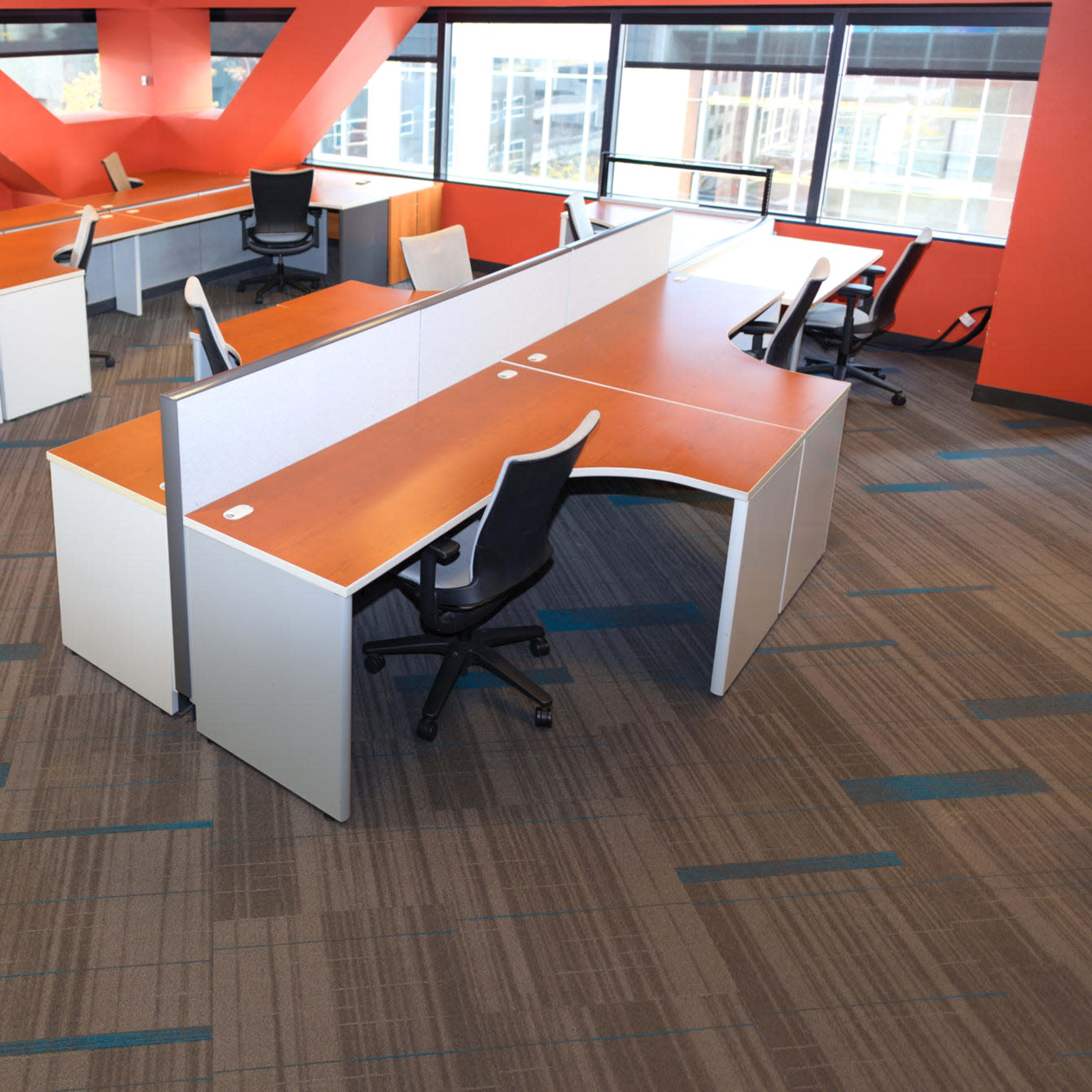 An office space features several modern desks with orange accents and ergonomic chairs arranged in a layout near large windows.