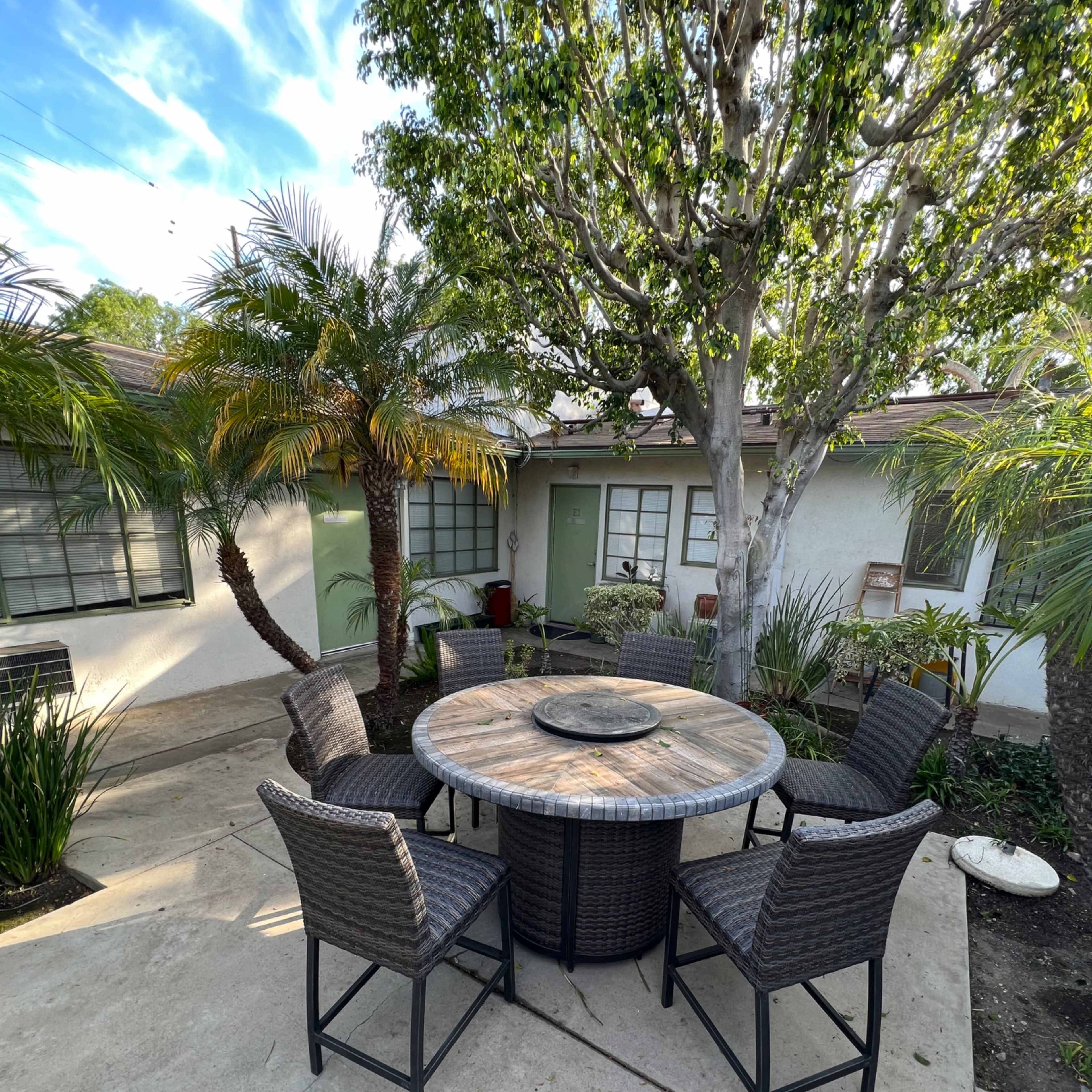 A round table with six high-backed chairs is placed in a landscaped courtyard surrounded by palm trees and foliage.