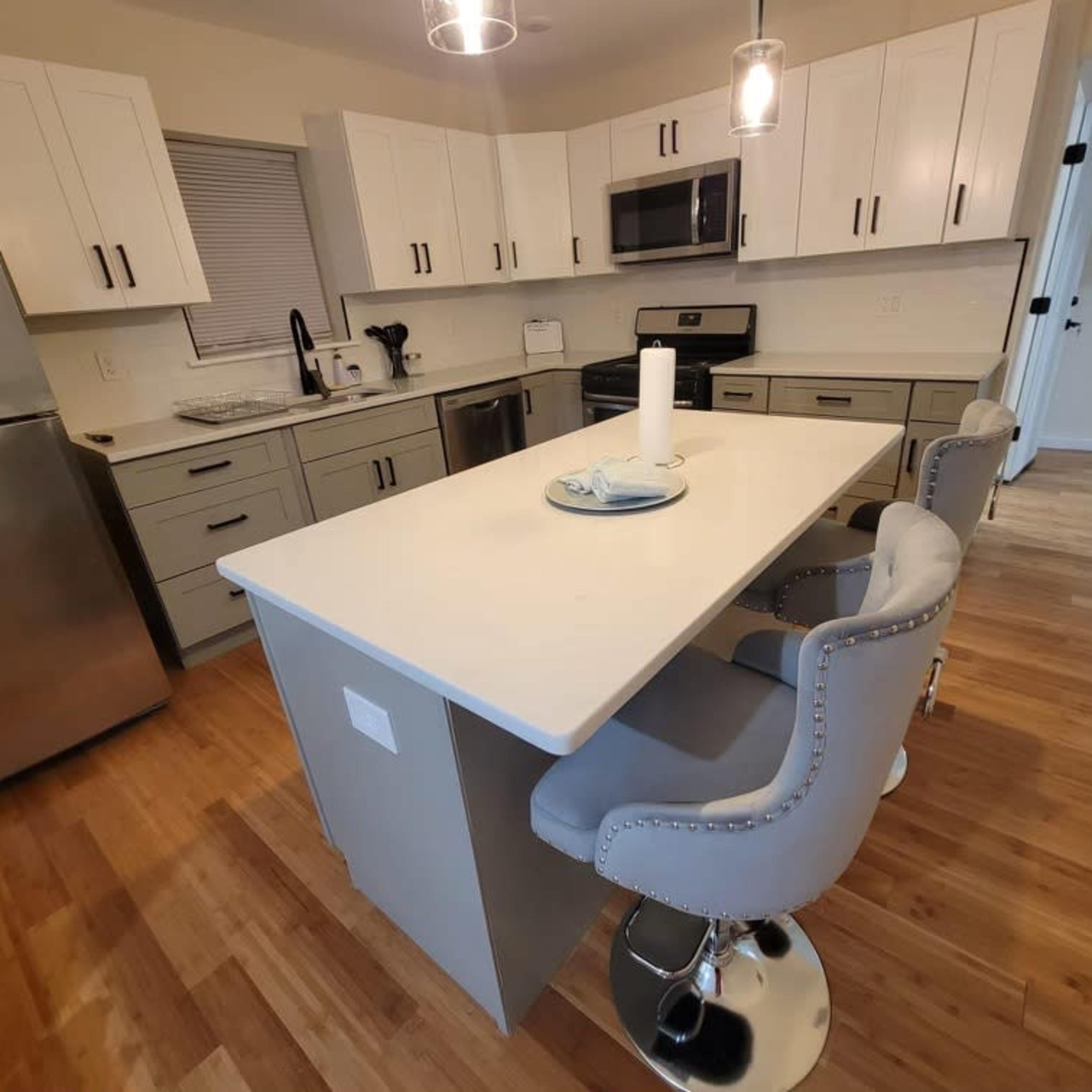 A modern kitchen with a central island, gray cabinetry, stainless steel appliances, and four upholstered counter stools.