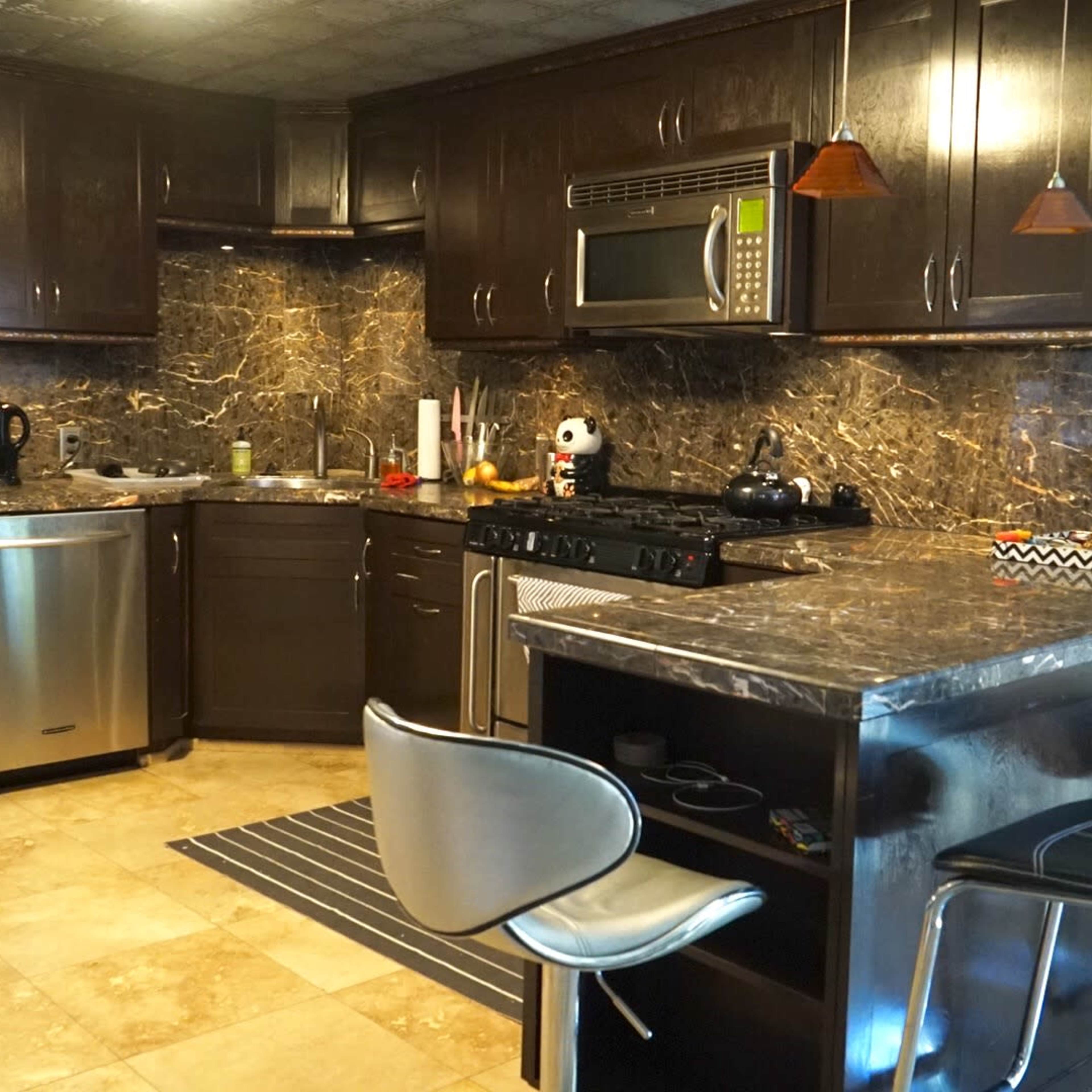 A modern kitchen with dark cabinetry, marble countertops, stainless steel appliances, and a bar with two chairs.