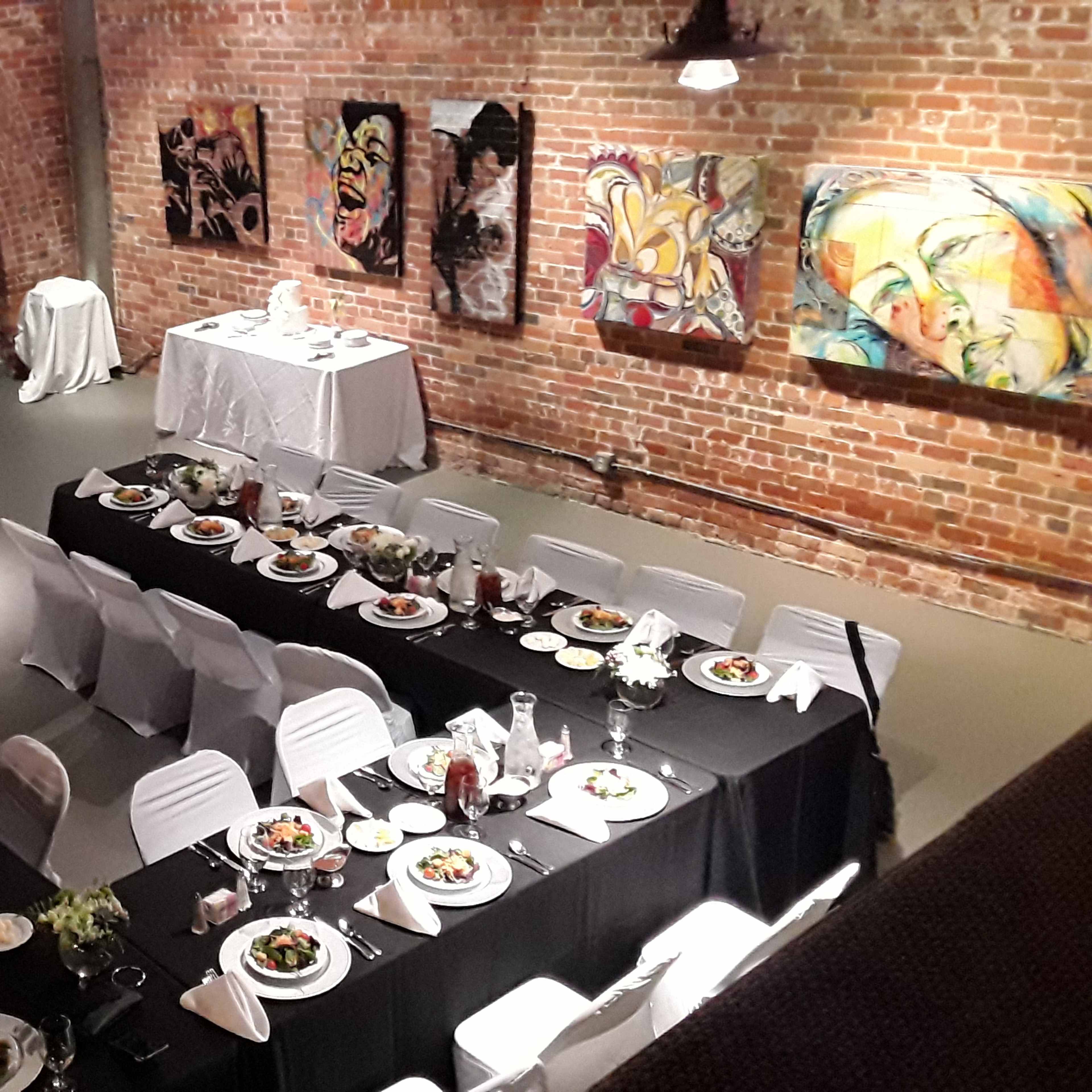 A well-set dining area with two long tables adorned with plates and floral centerpieces, surrounded by brick walls and colorful artwork.